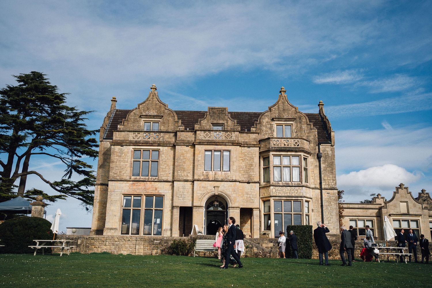 Panoramic view of a grand stone manor house with Tudor-style architecture under blue skies.