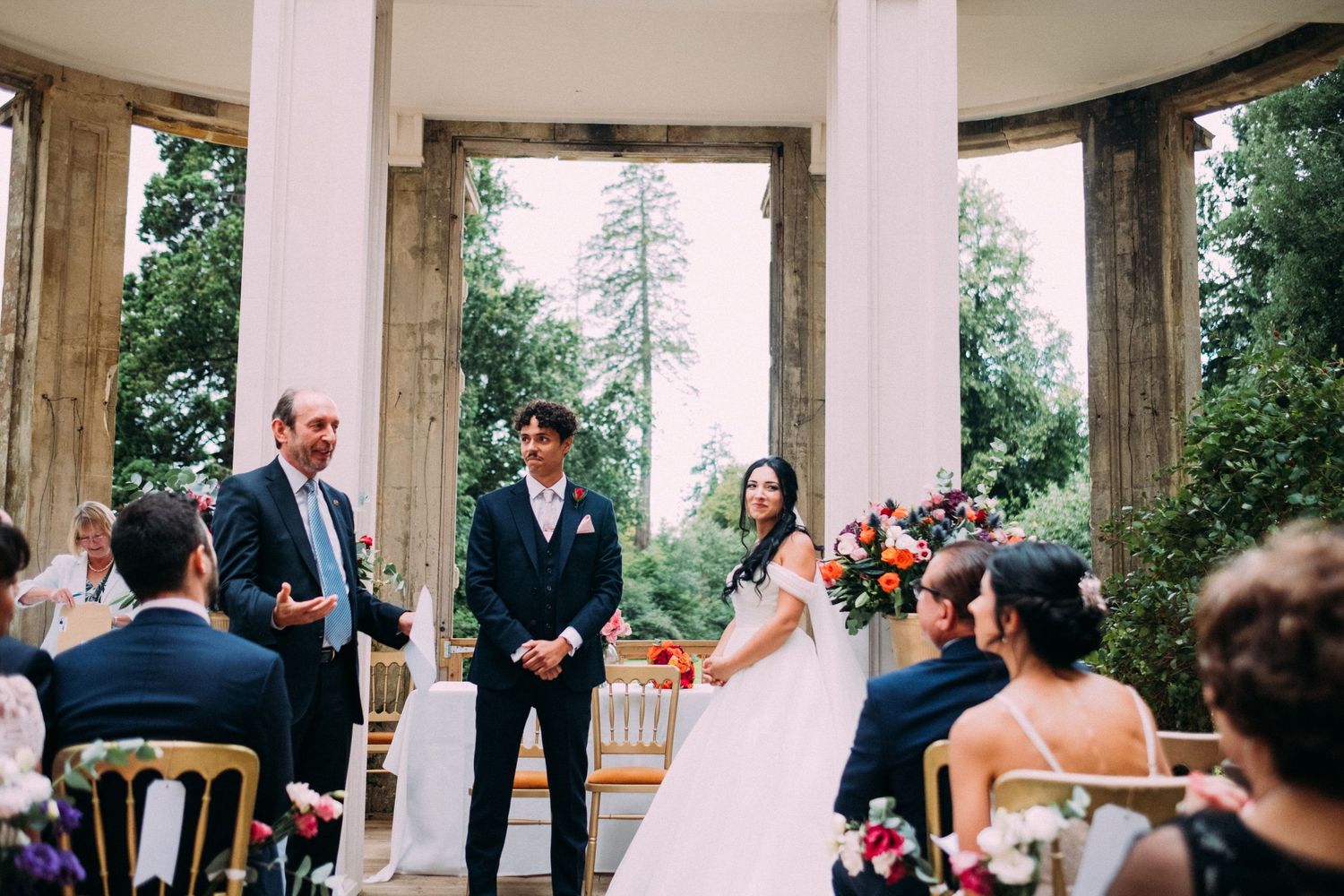 Wedding ceremony taking place in an elegant outdoor gazebo with white columns surrounded by greenery.
