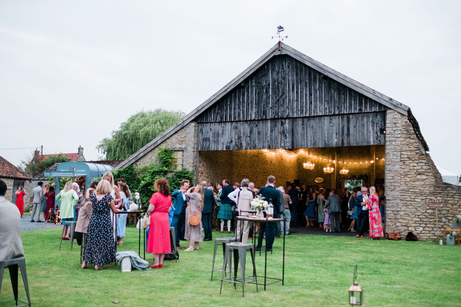 Outdoor event venue with rustic barn buildings and guests gathering on a grassy lawn during a celebration.