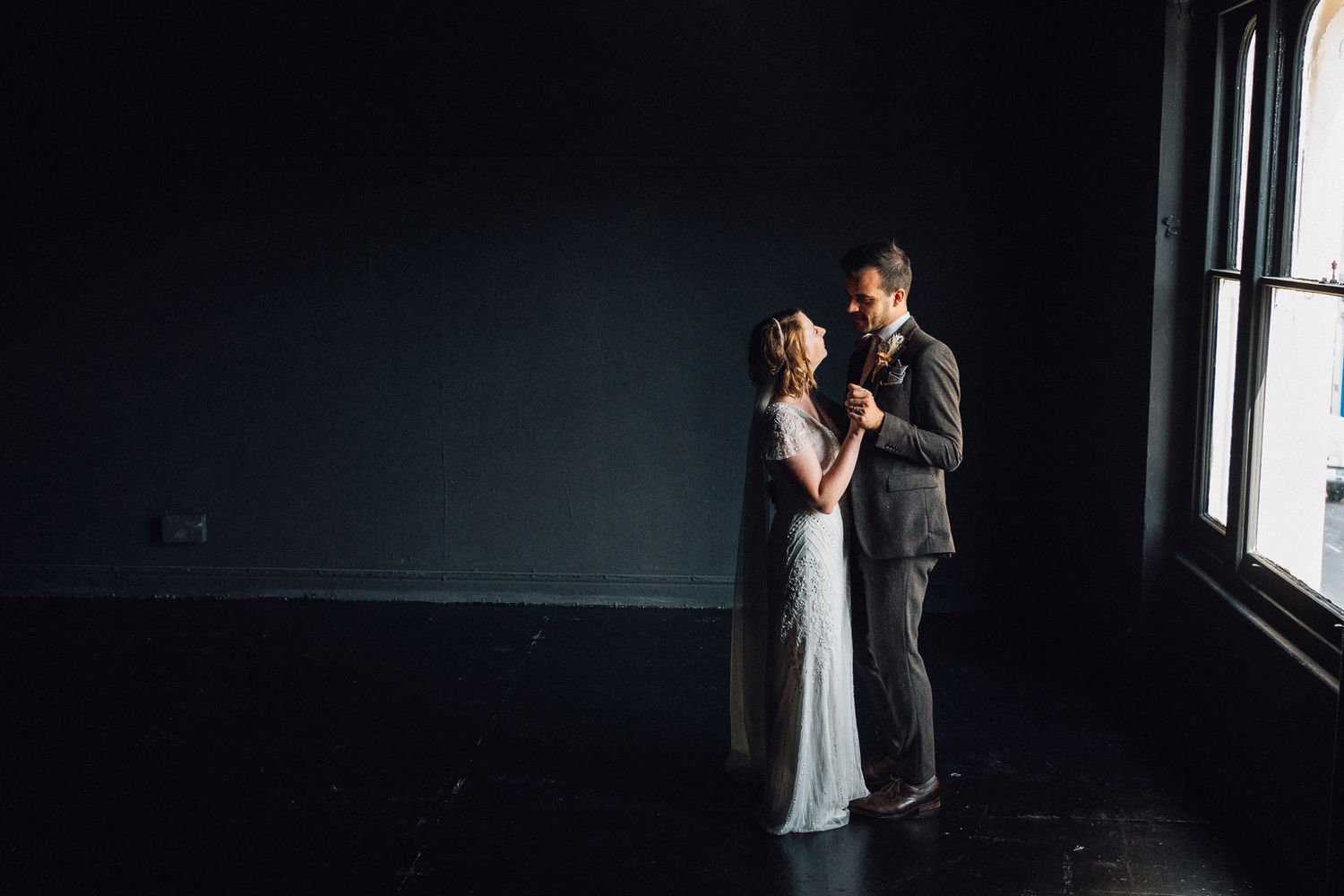 A romantic couple in formal attire shares an intimate moment in a dimly lit room by a window.