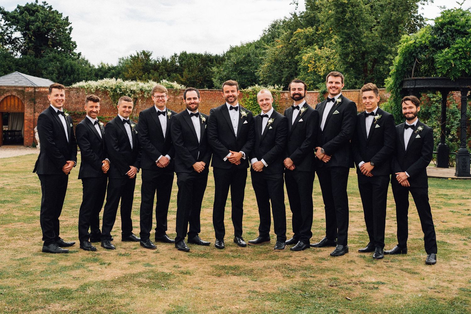 A wedding party in black tuxedos and bow ties stands in a line on grass with trees in the background.