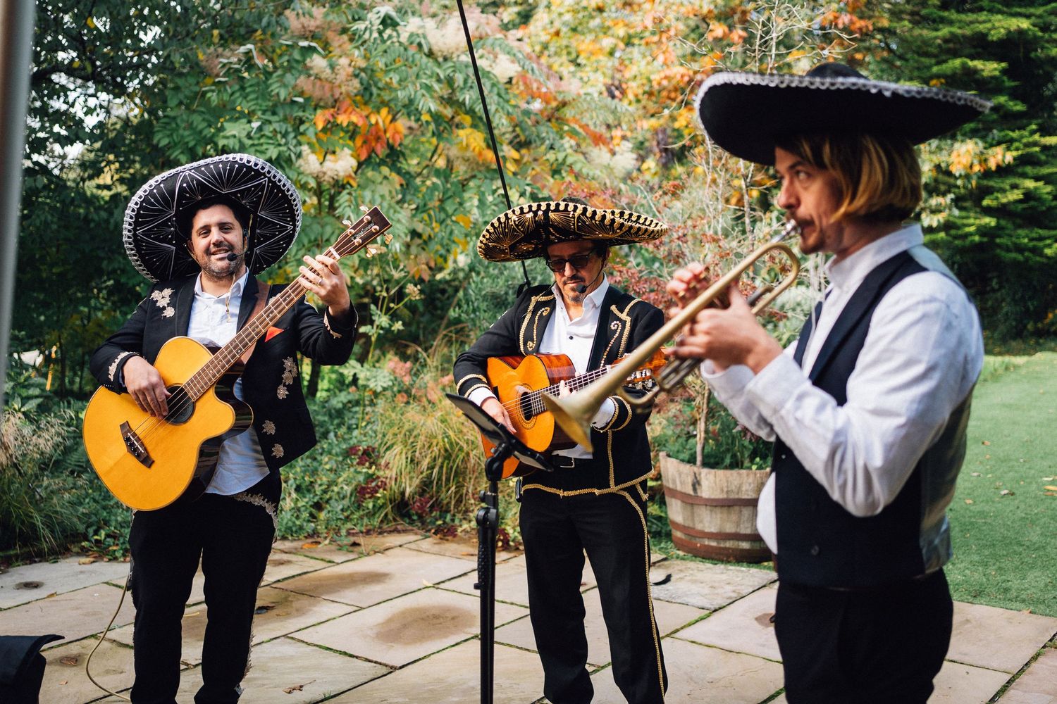 Three mariachi musicians in traditional attire perform outdoors with guitars and trumpet in a garden setting.