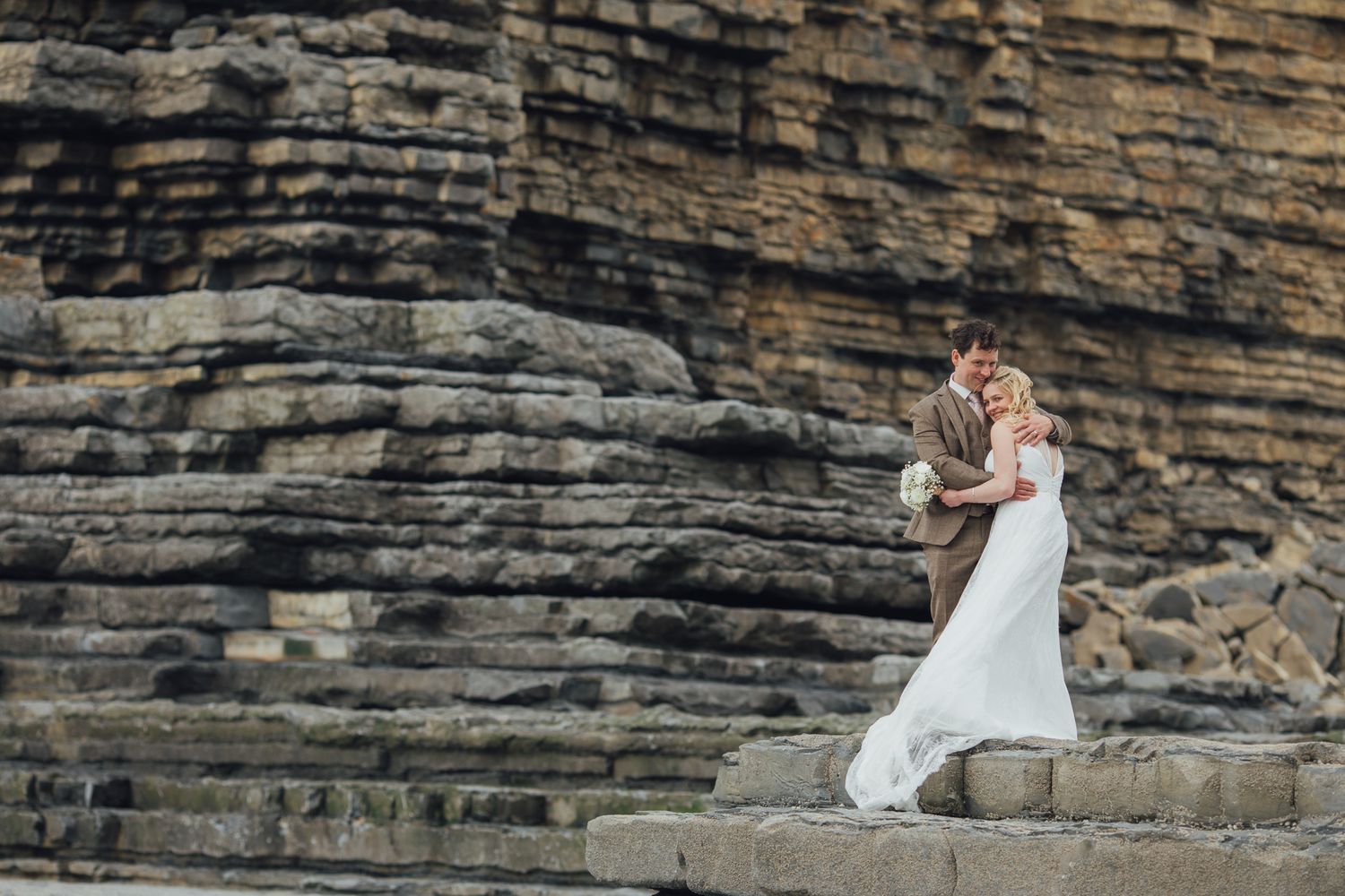 A wedding couple stands on stone steps against dramatic layered rock formations.