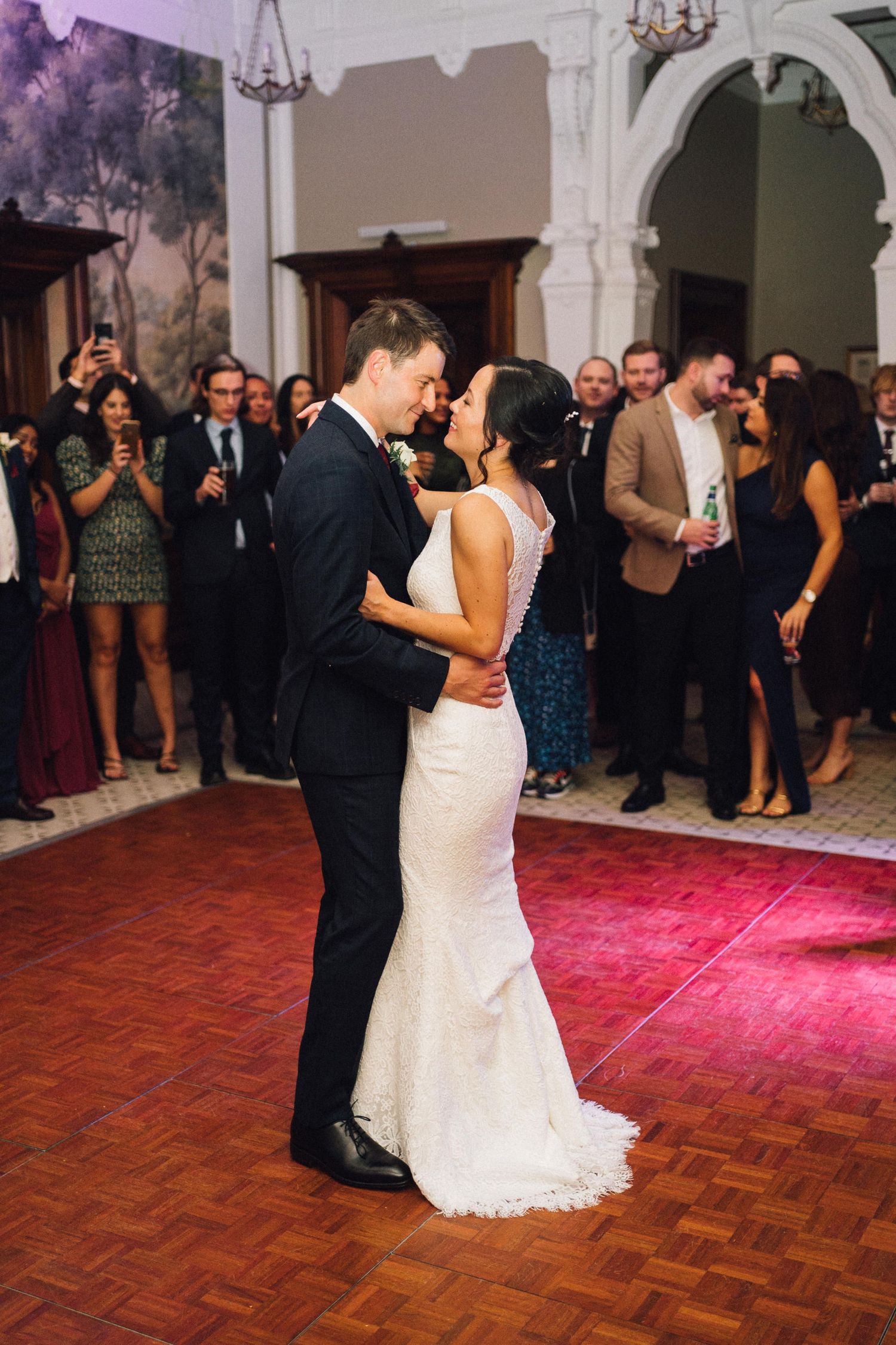 Couple dancing together at an elegant wedding reception on a parquet dance floor with guests watching.