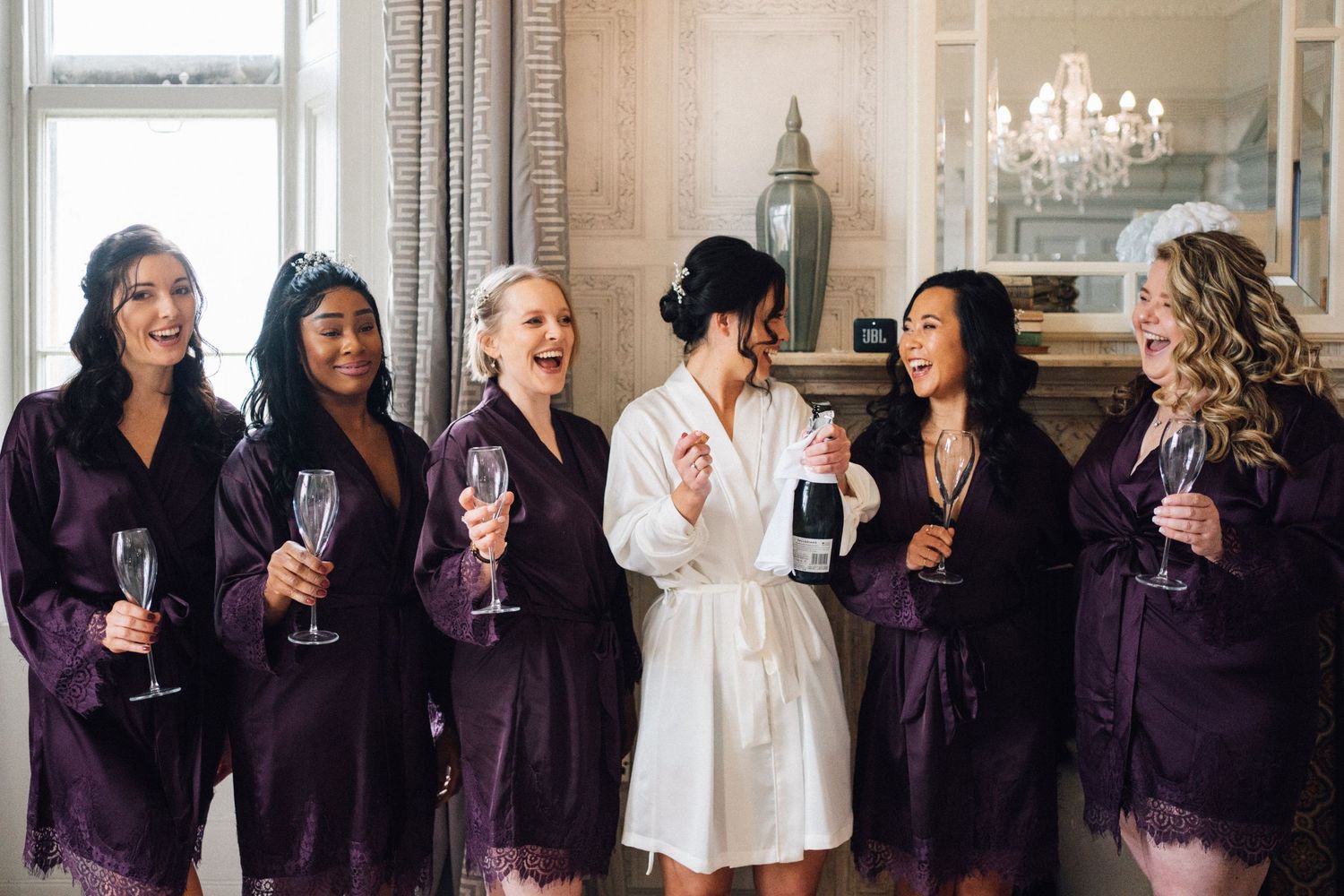 Wedding party wearing purple and white robes celebrating with champagne in a bright room.
