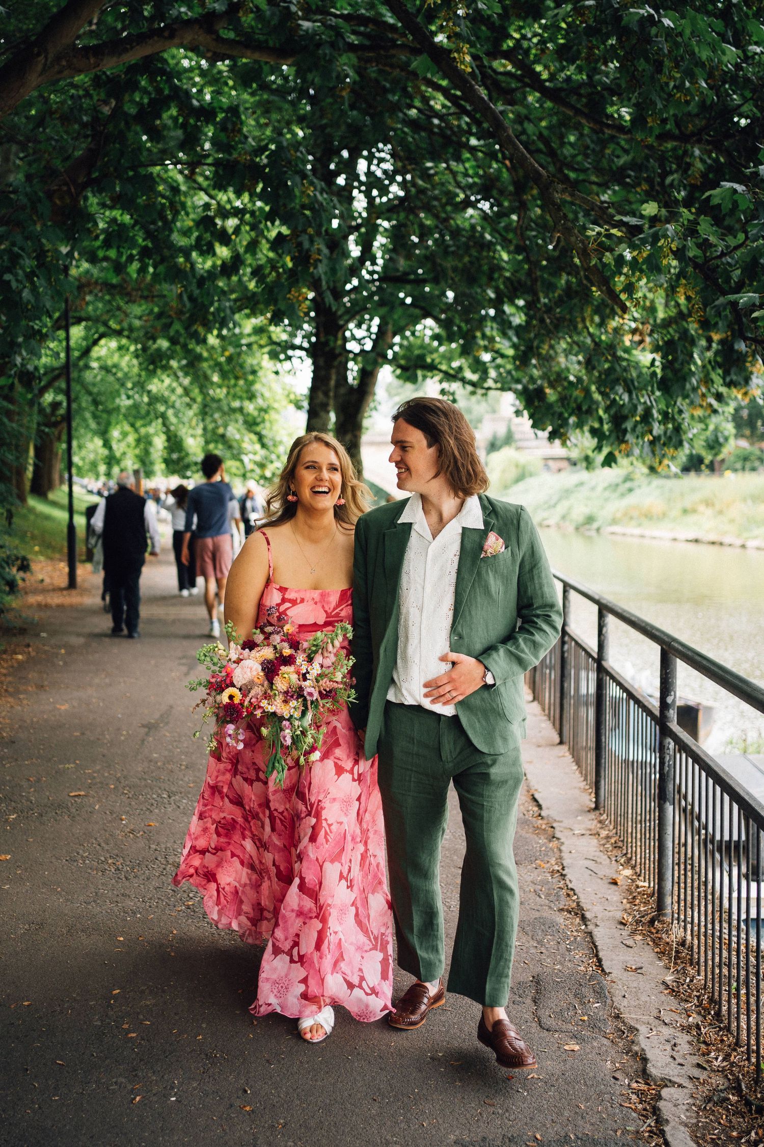 A couple walks together on a tree-lined path, one wearing a pink floral dress and another in a green suit.