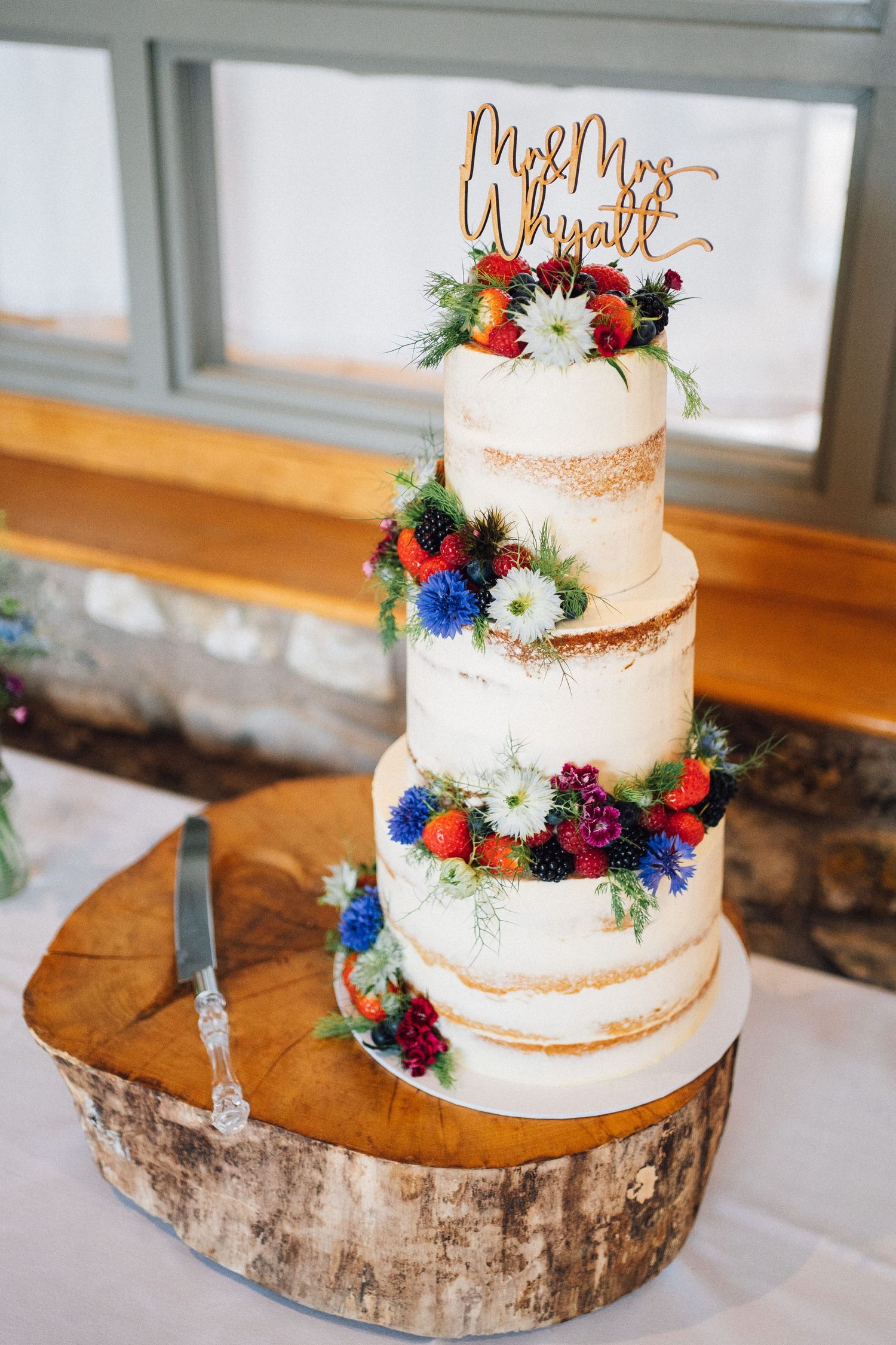 Four-tier semi-naked wedding cake decorated with colorful flowers on rustic wood slice stand.
