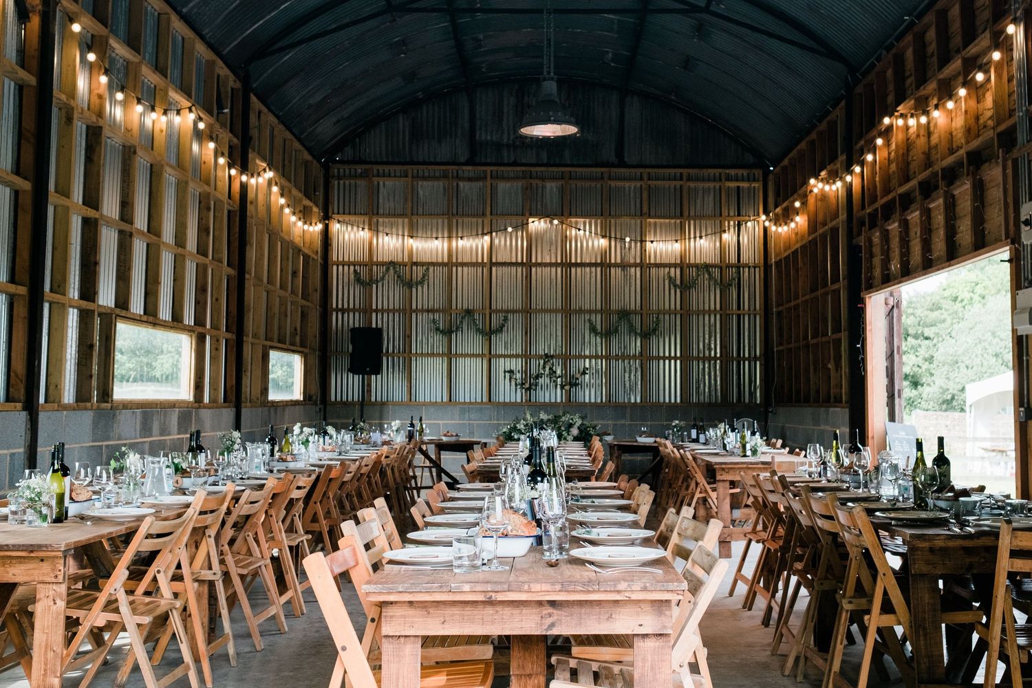 Long wooden banquet tables and chairs set up in a rustic barn venue with string lights hanging overhead.