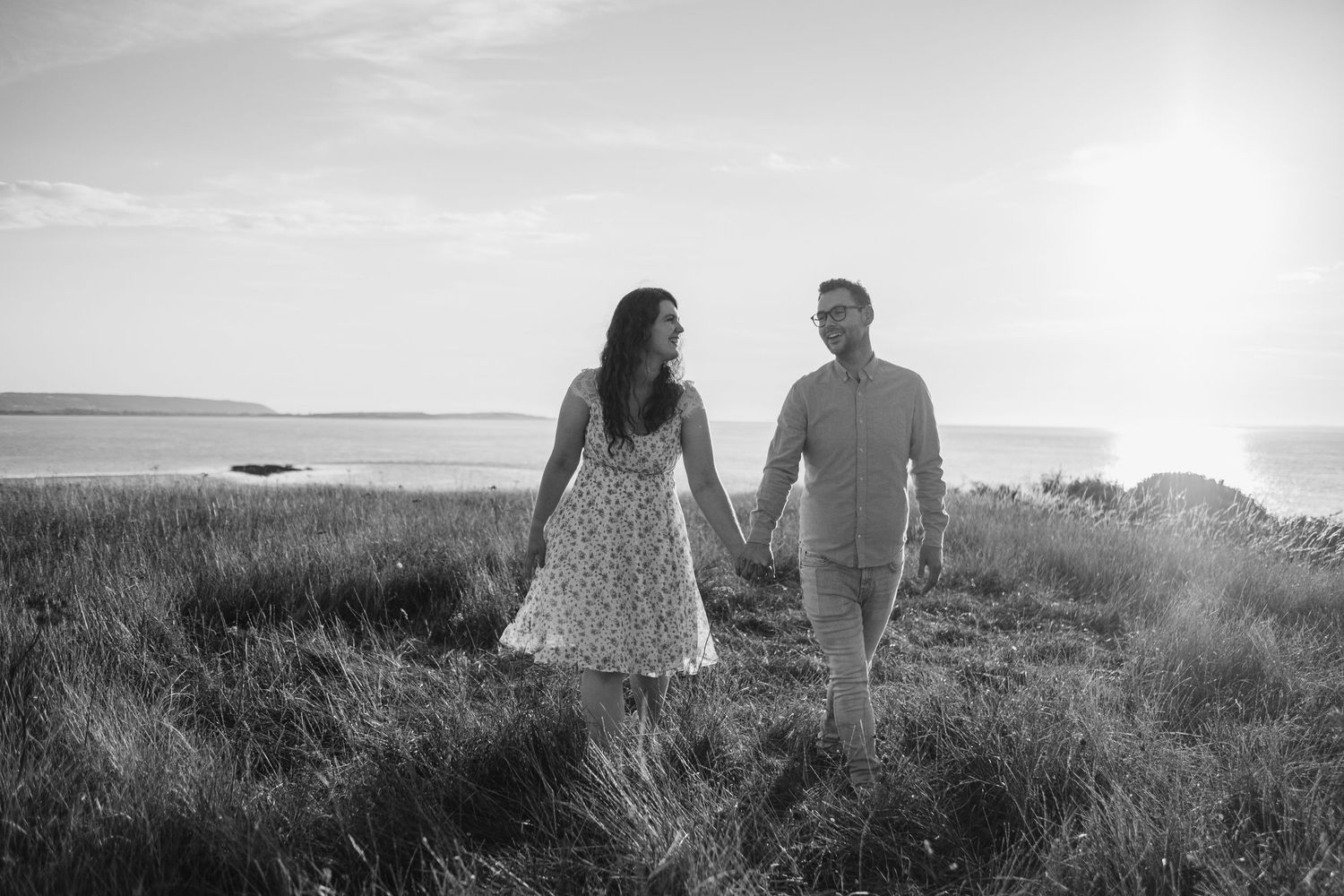 Romantic couple walking together through tall grass near the ocean at sunset in black and white.