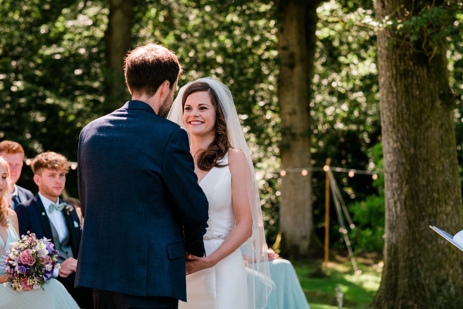 Outdoor wedding ceremony taking place under tall trees with natural lighting.