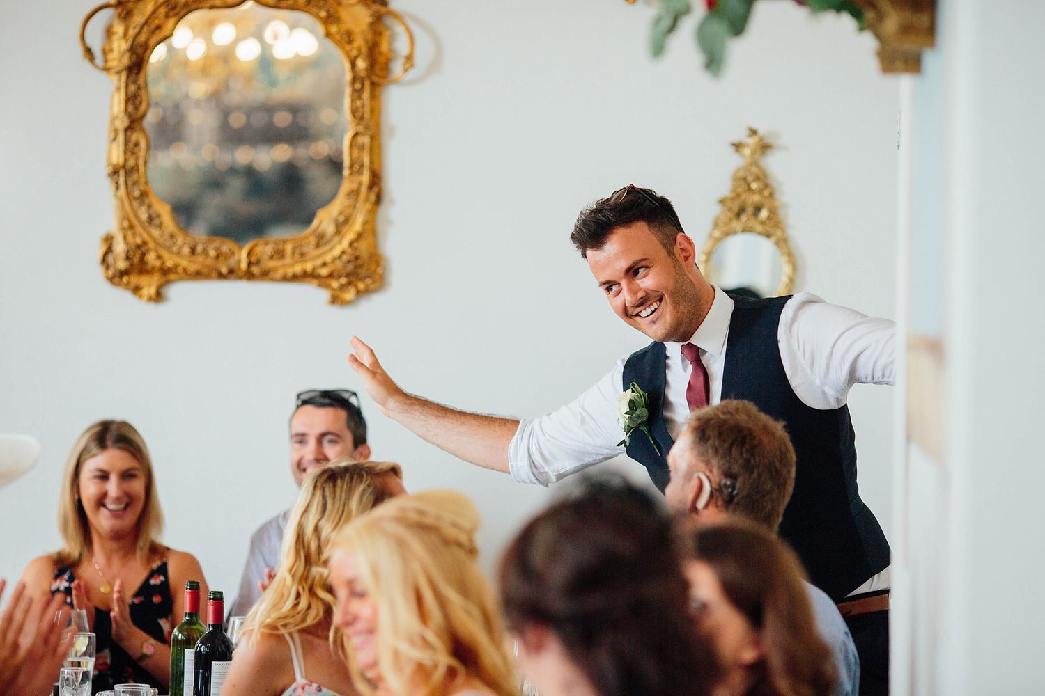 A speaker in formal attire gestures enthusiastically while delivering a speech during a wedding reception.