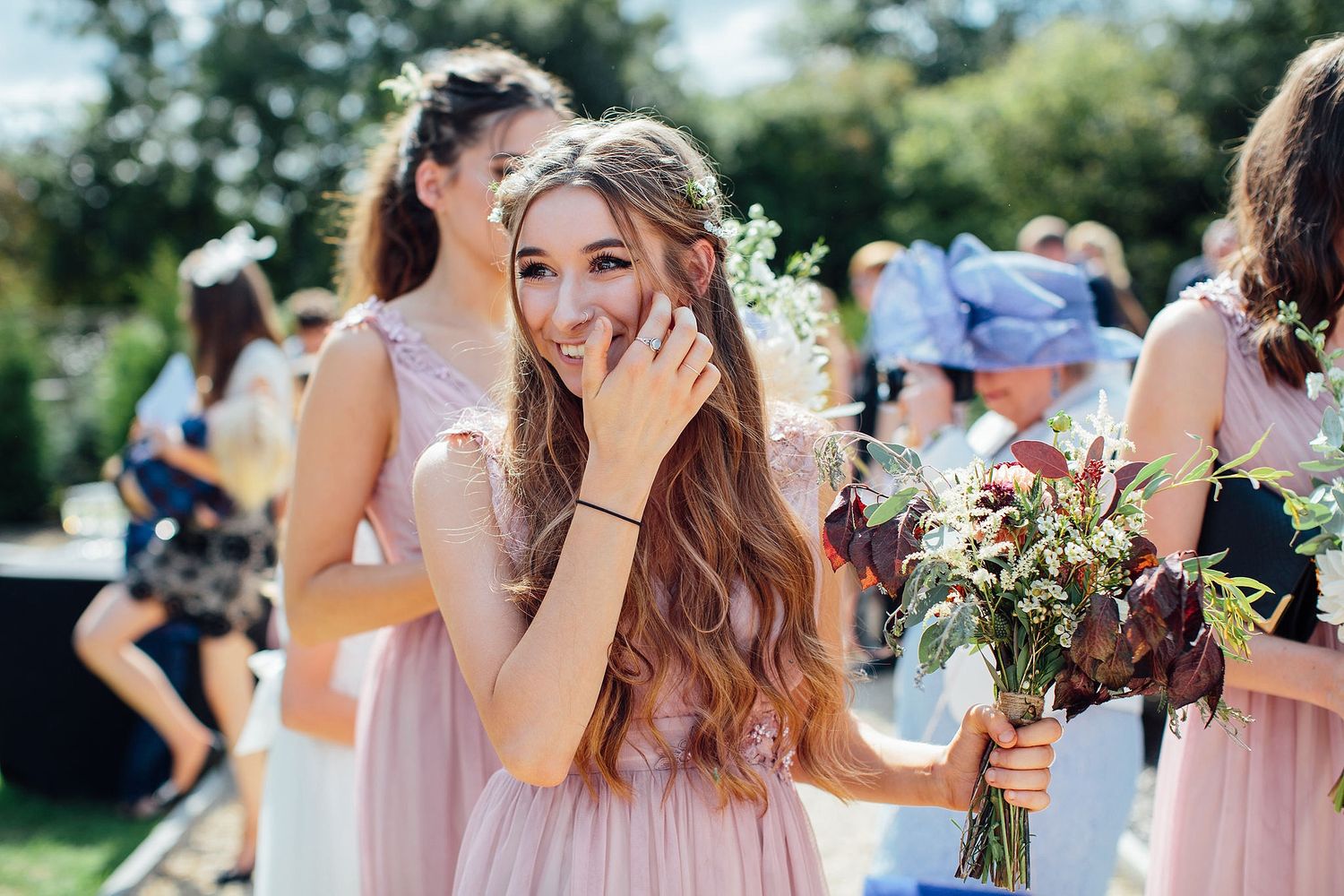 Wedding guests in pink dresses holding bouquets during an outdoor summer celebration.