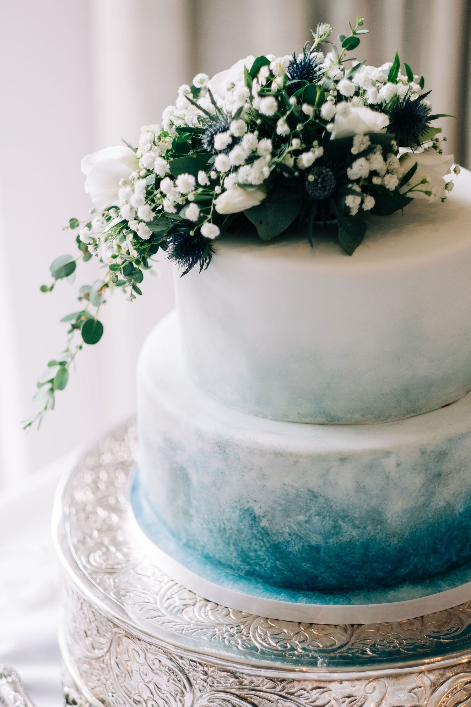 Blue ombre wedding cake decorated with white flowers and greenery on a silver cake stand.