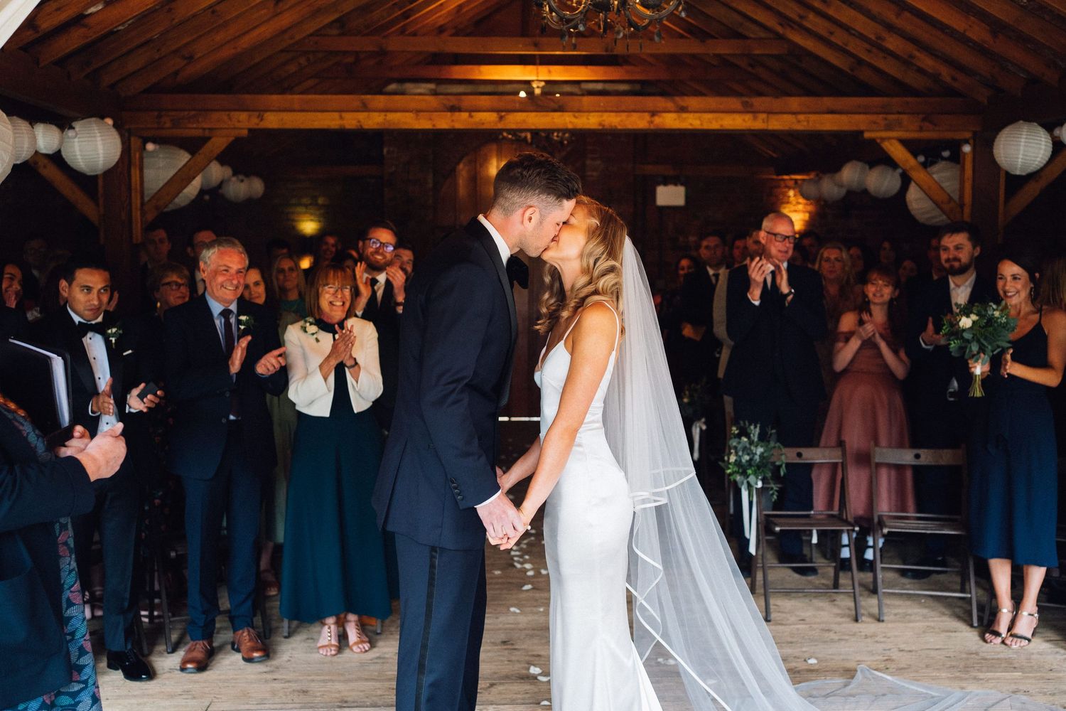Wedding reception guests dancing and celebrating under a rustic wooden barn ceiling with hanging lanterns.