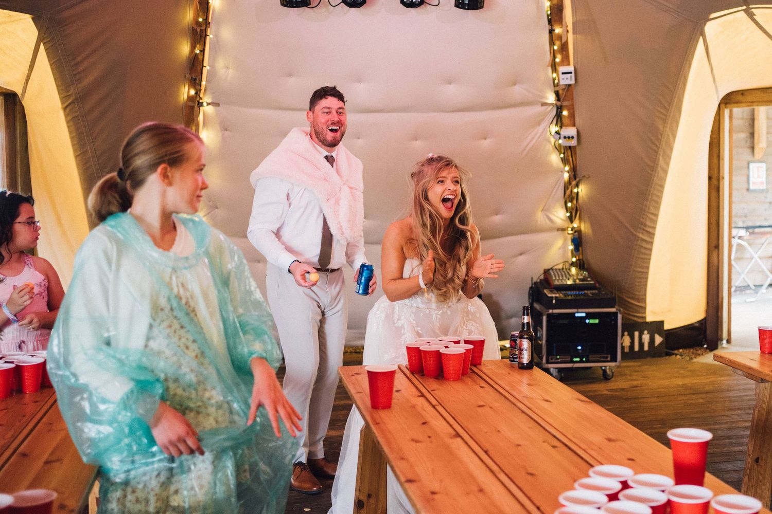 Party guests in colorful rain ponchos dancing and laughing inside a tent with string lights.