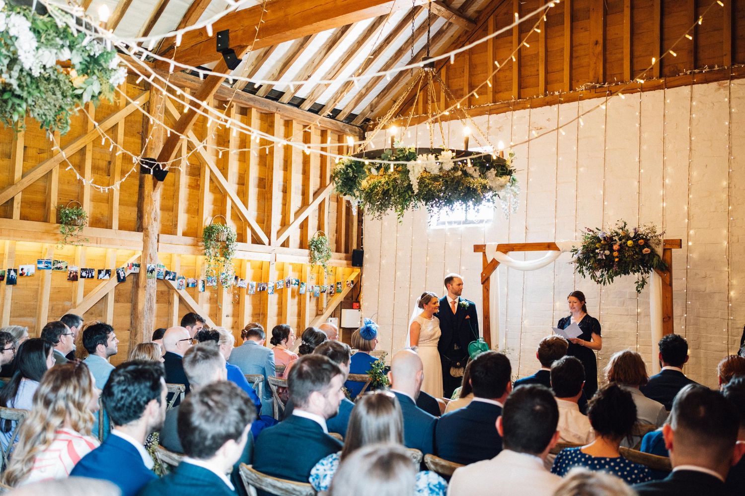 Wide view of a barn wedding ceremony with exposed wooden beams and string lights.