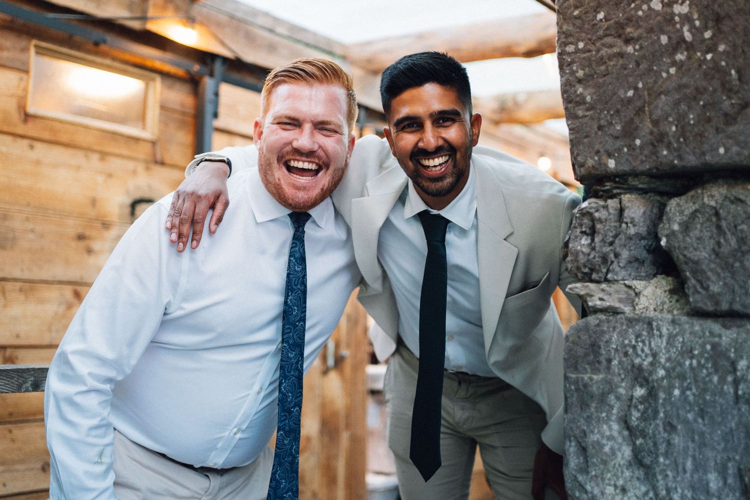 Two people in formal attire share a joyful moment while standing against a rustic wooden wall.