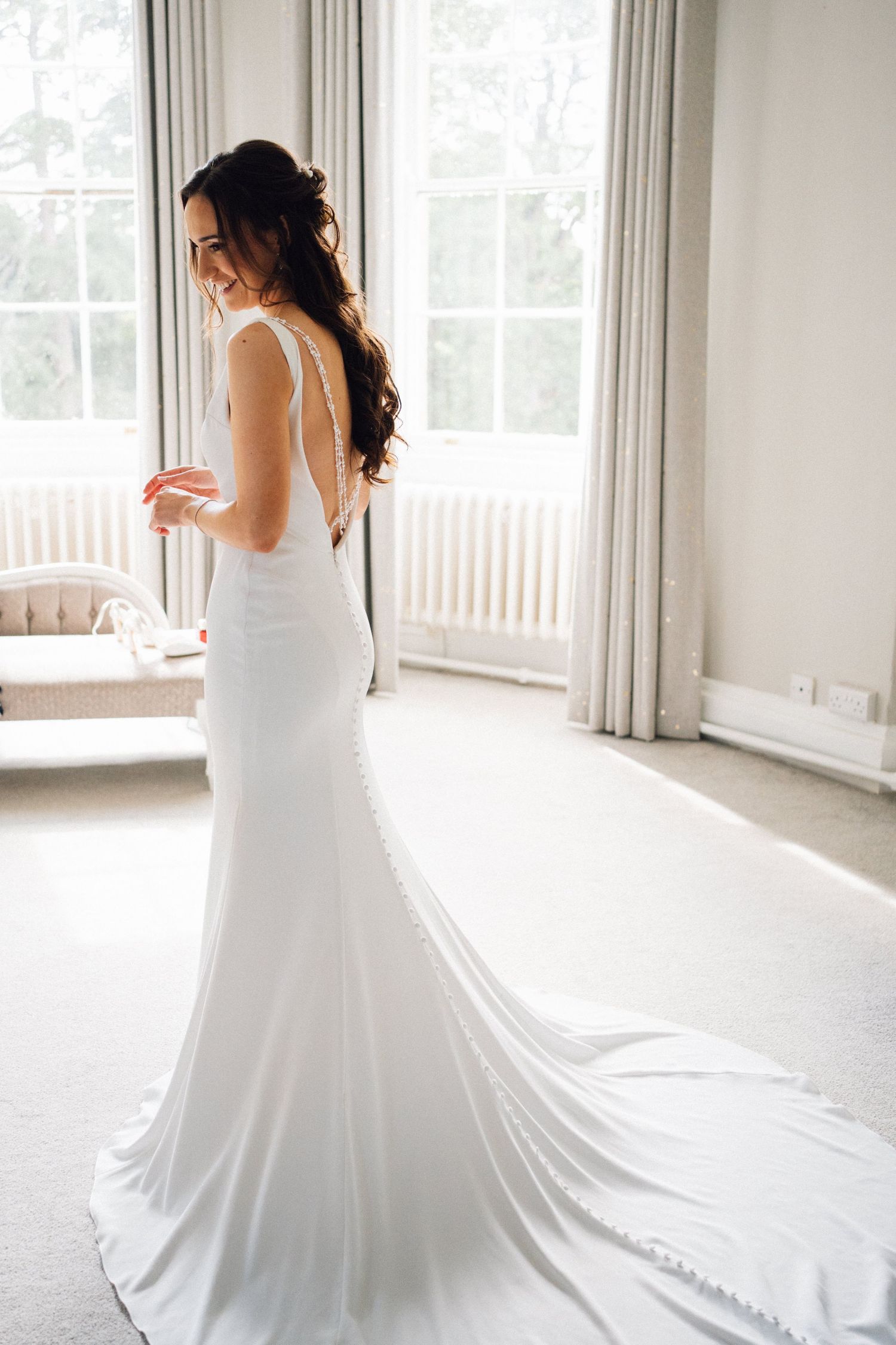 A bride in a flowing white wedding dress twirls elegantly in a bright sunlit room with floor-to-ceiling windows.