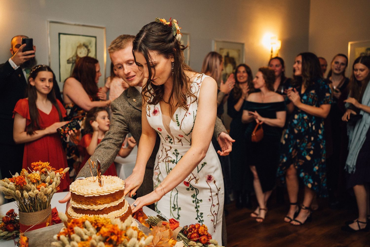 Couple cuts their wedding cake together at reception while guests watch in background.