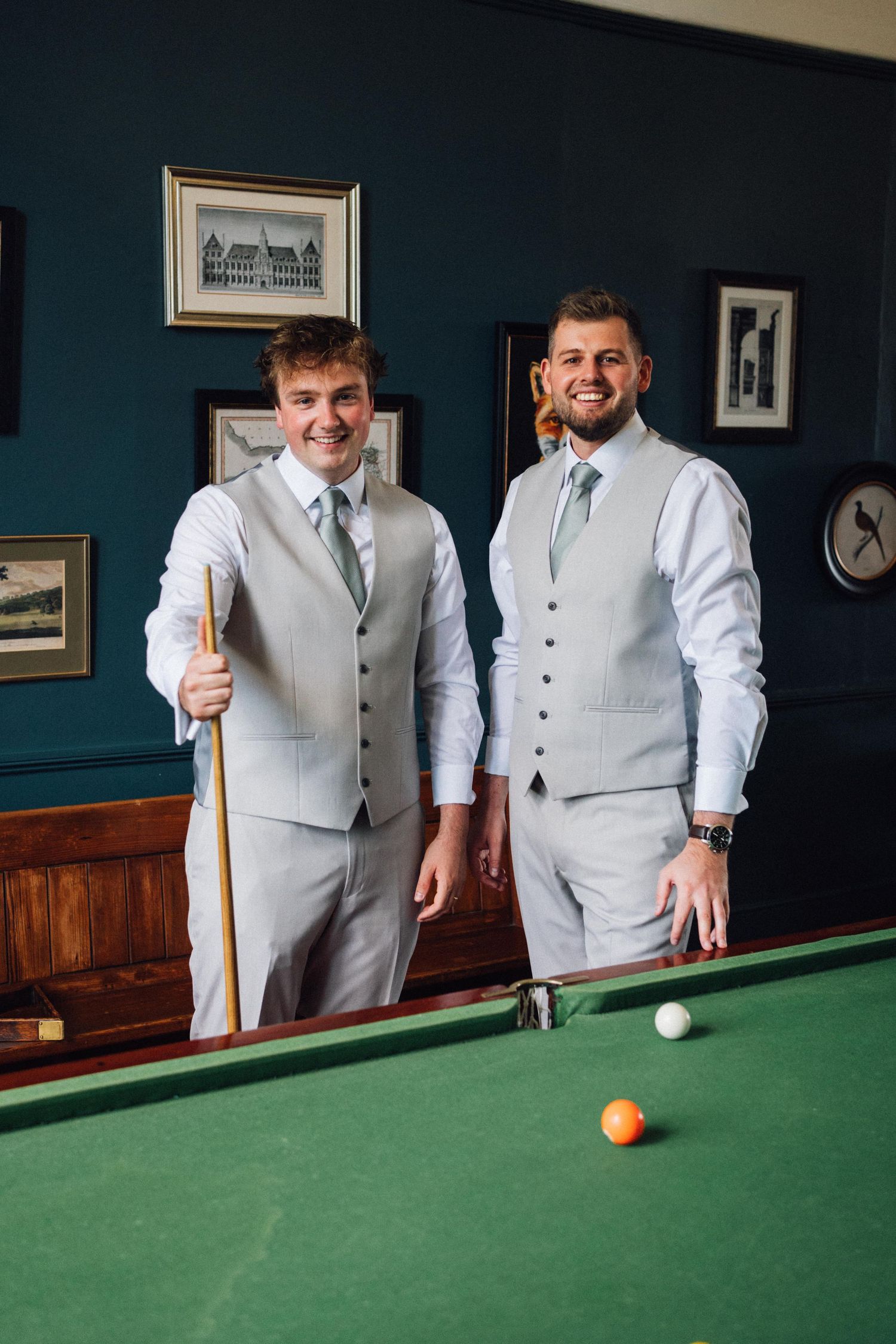 Groomsmen in light gray vests playing pool on green felt table in vintage room with dark walls.