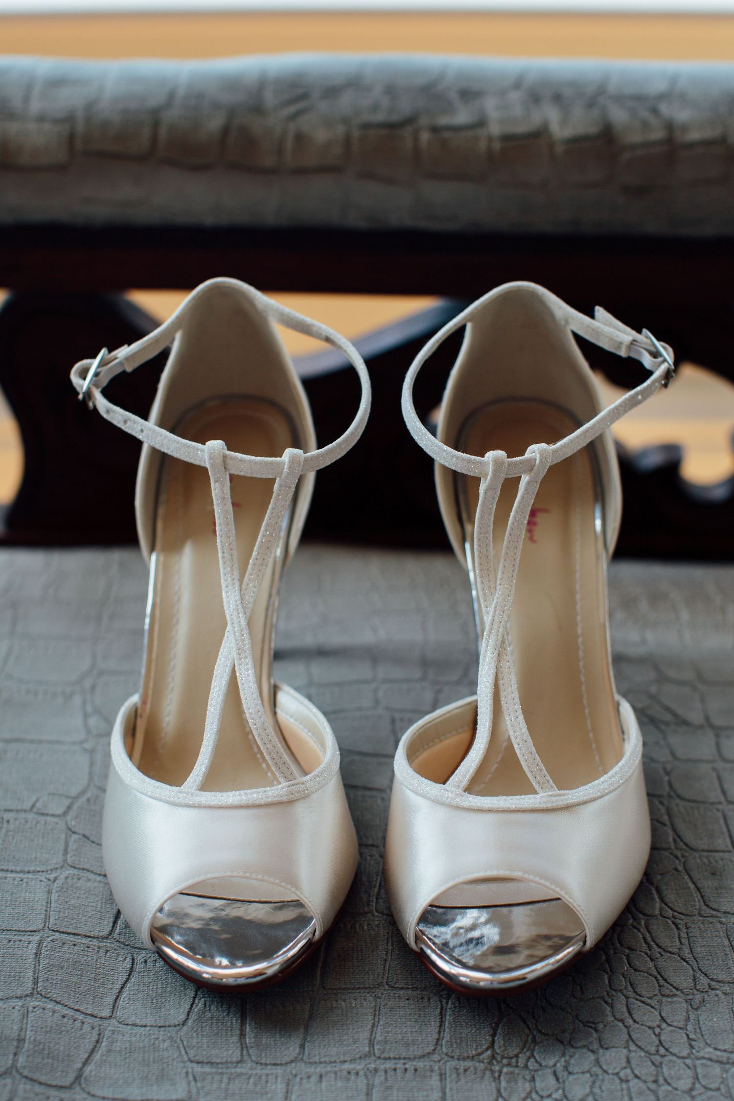 Elegant white satin T-strap bridal shoes with peep toe detail on textured surface.
