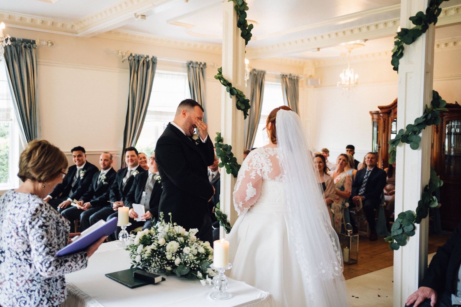 Wedding ceremony guests gather in an elegant reception hall decorated with white floral arrangements and turquoise curtains.