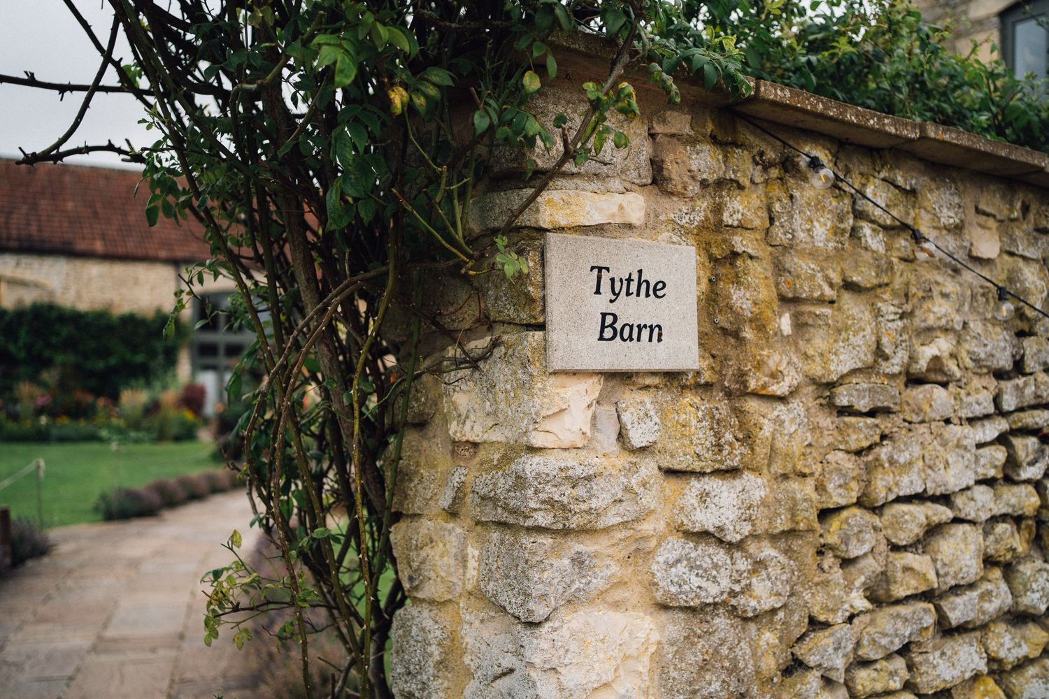 Stone wall sign reading Tythe Barn covered with climbing plants at a rustic countryside venue.