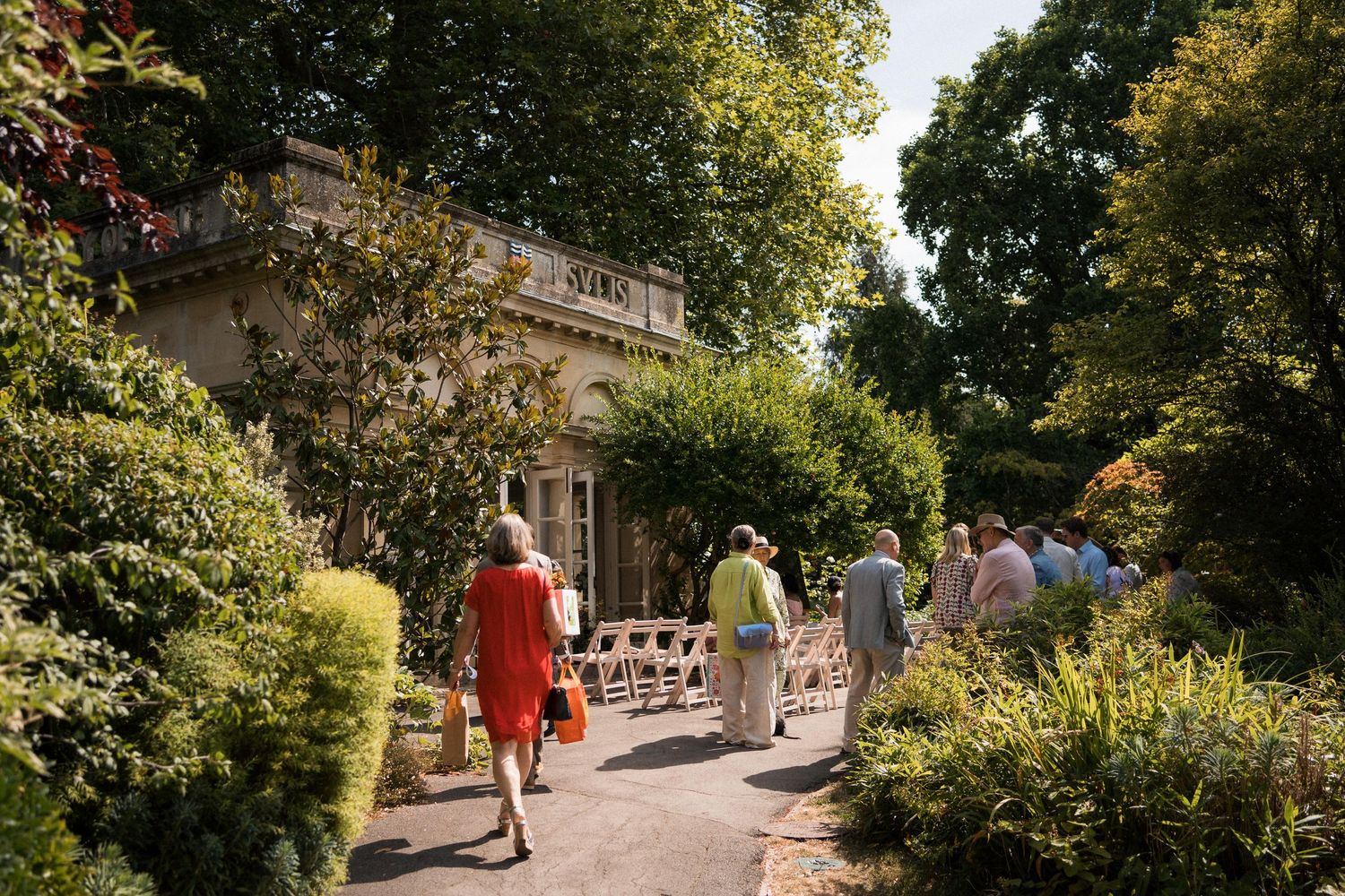 Guests walk along a sunlit garden path toward an elegant historic building surrounded by lush green trees and shrubs.