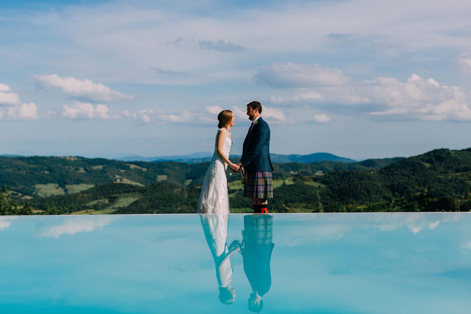 A couple stands together on a balcony overlooking a stunning turquoise infinity pool with mountain views in the background.