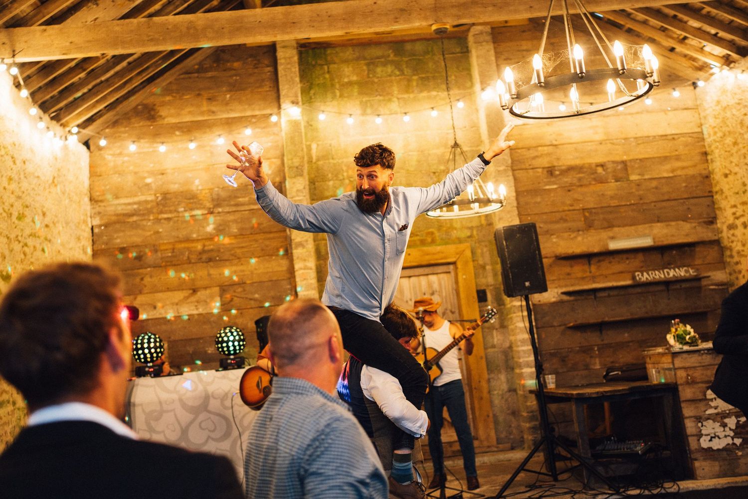 Wedding guests dancing and celebrating in a rustic stone-walled venue with wooden beams and string lights.