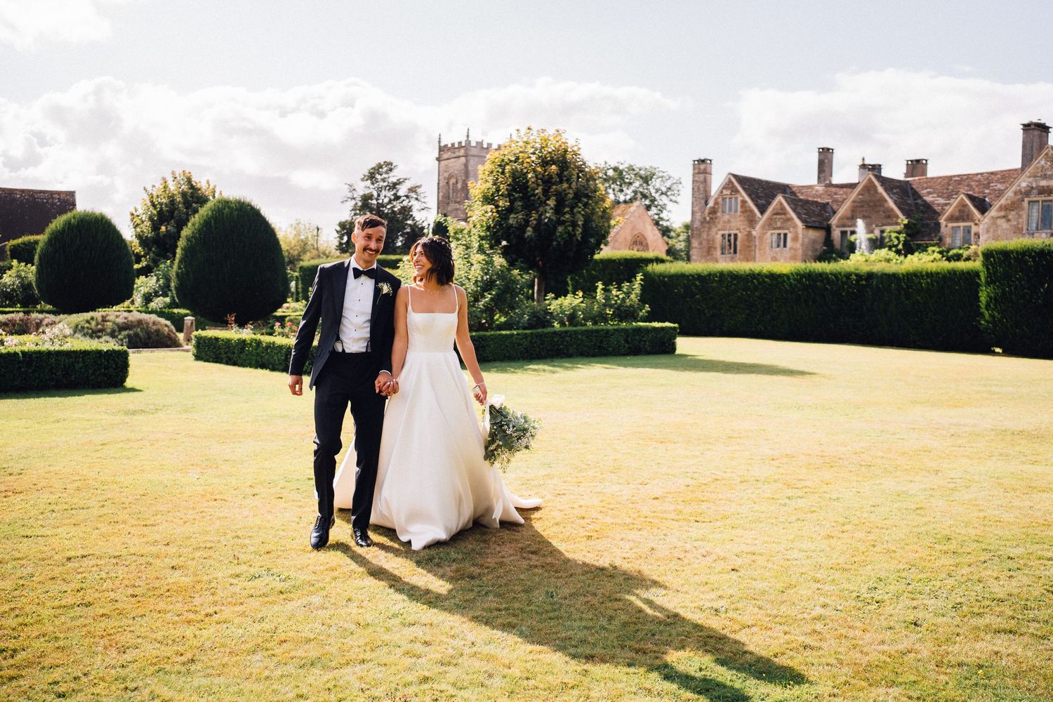Wedding couple walking hand in hand across a sunlit meadow with buildings in the distance.