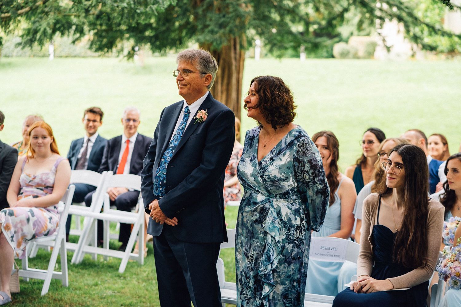 Outdoor garden wedding ceremony with guests seated on white chairs under trees.