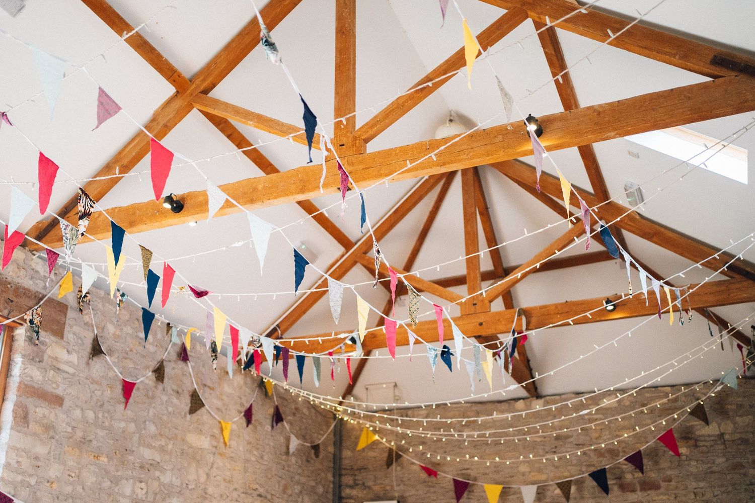 Exposed wooden beams decorated with string lights and colorful triangle bunting in a rustic venue.