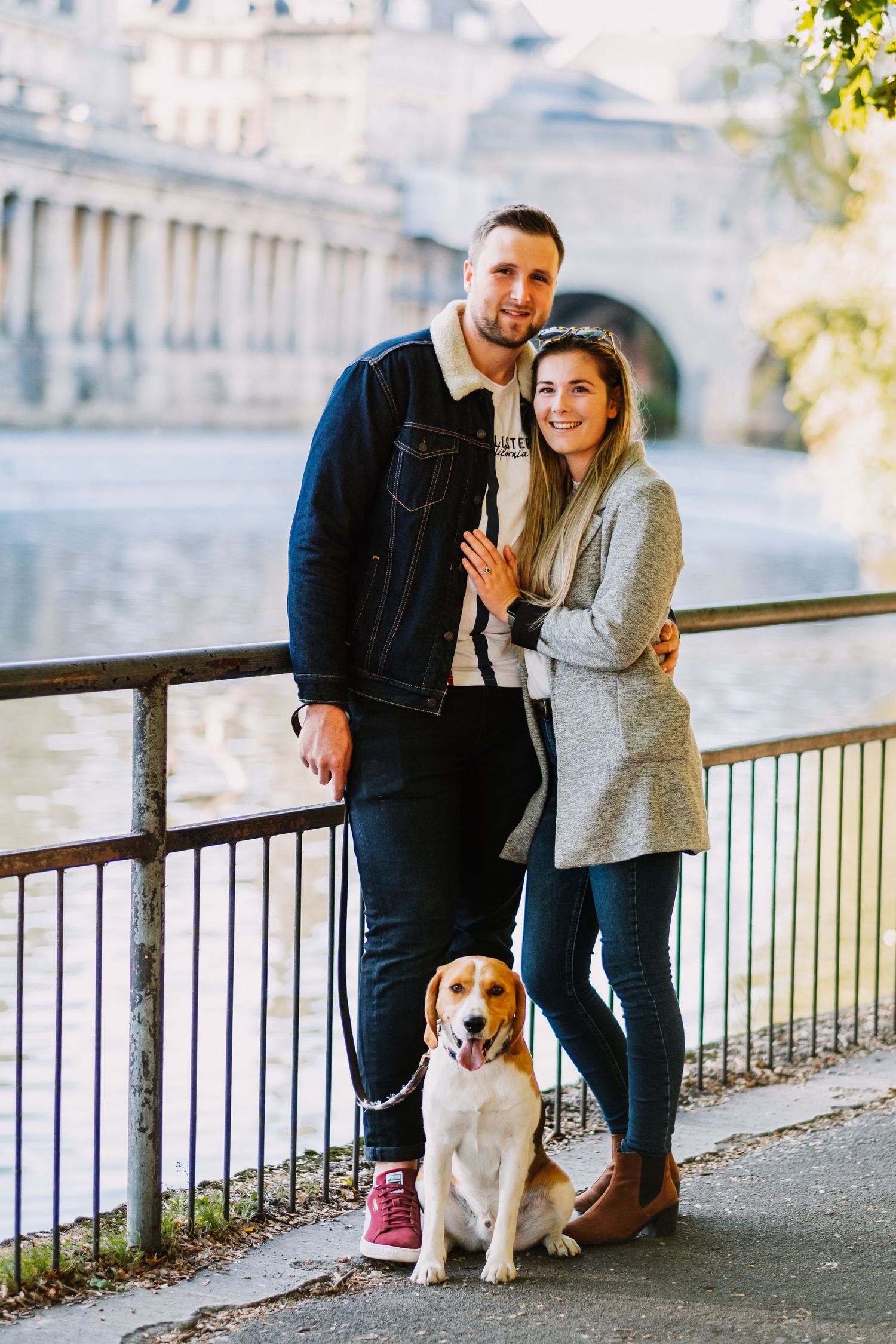 A couple poses with their beagle dog along a riverside railing.