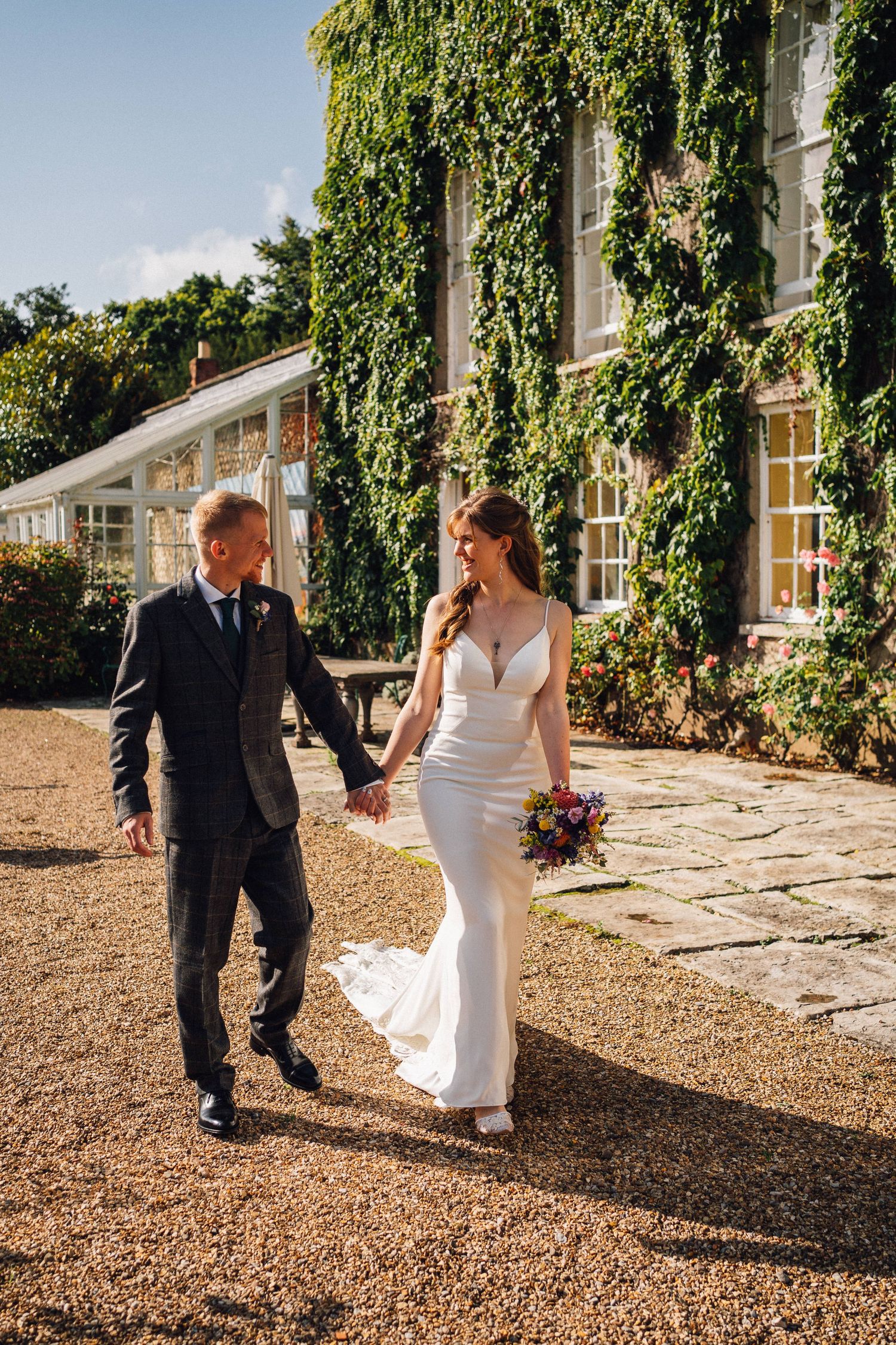 Couple walking along gravel path beside ivy-covered building during wedding photoshoot.