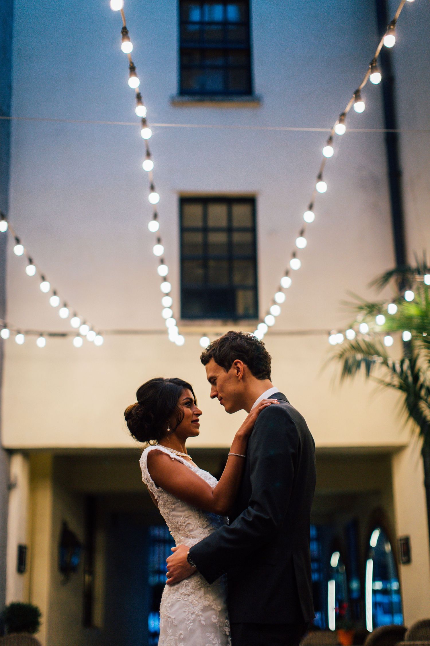 A couple shares an intimate moment beneath glowing string lights in an outdoor courtyard.