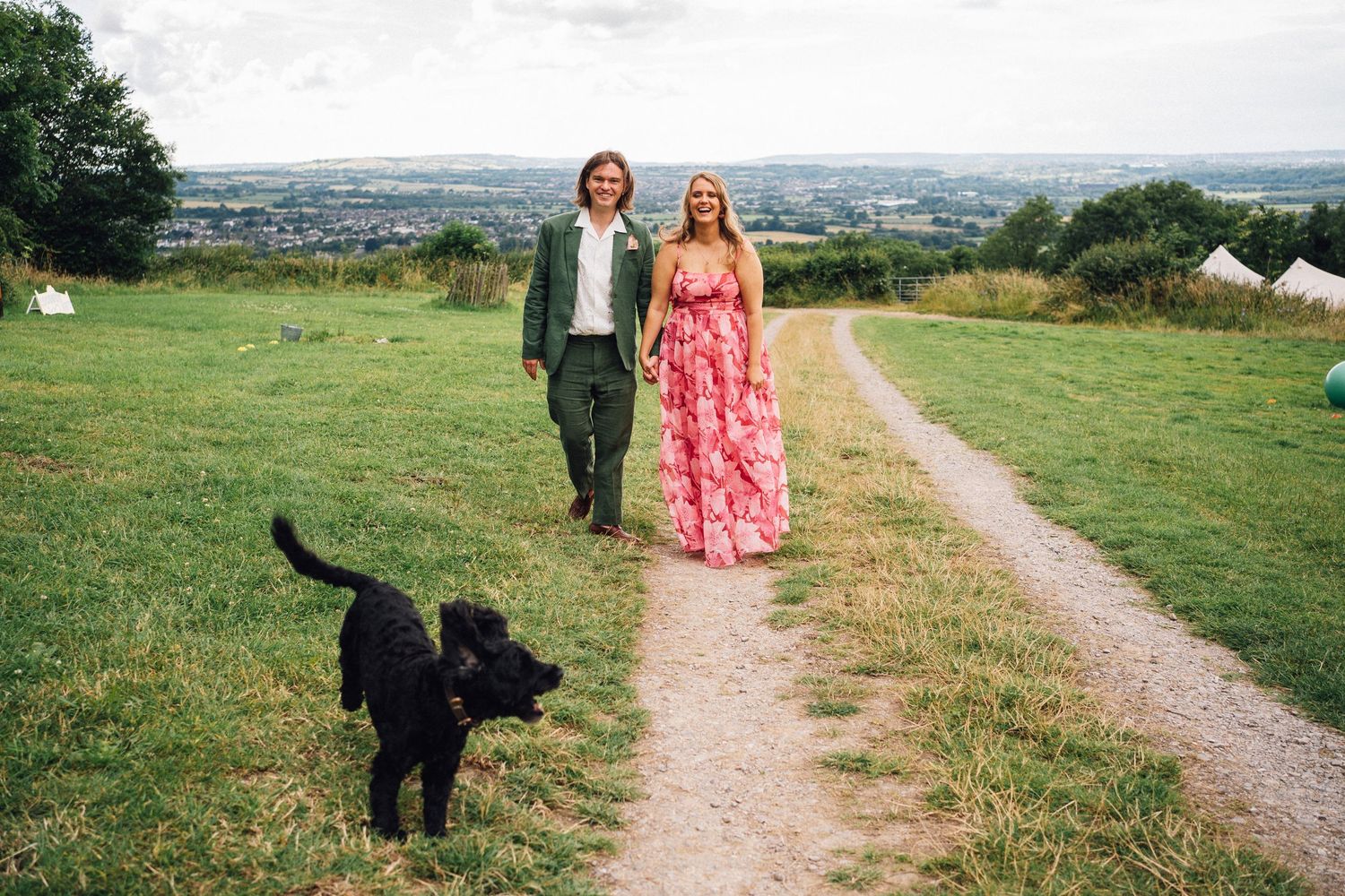 A scenic countryside path with green fields and a black dog walking in the distance.