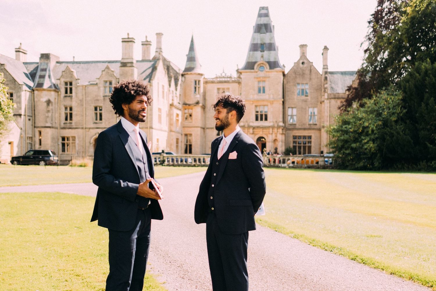 Two figures in formal attire standing together on the grounds of a historic mansion.