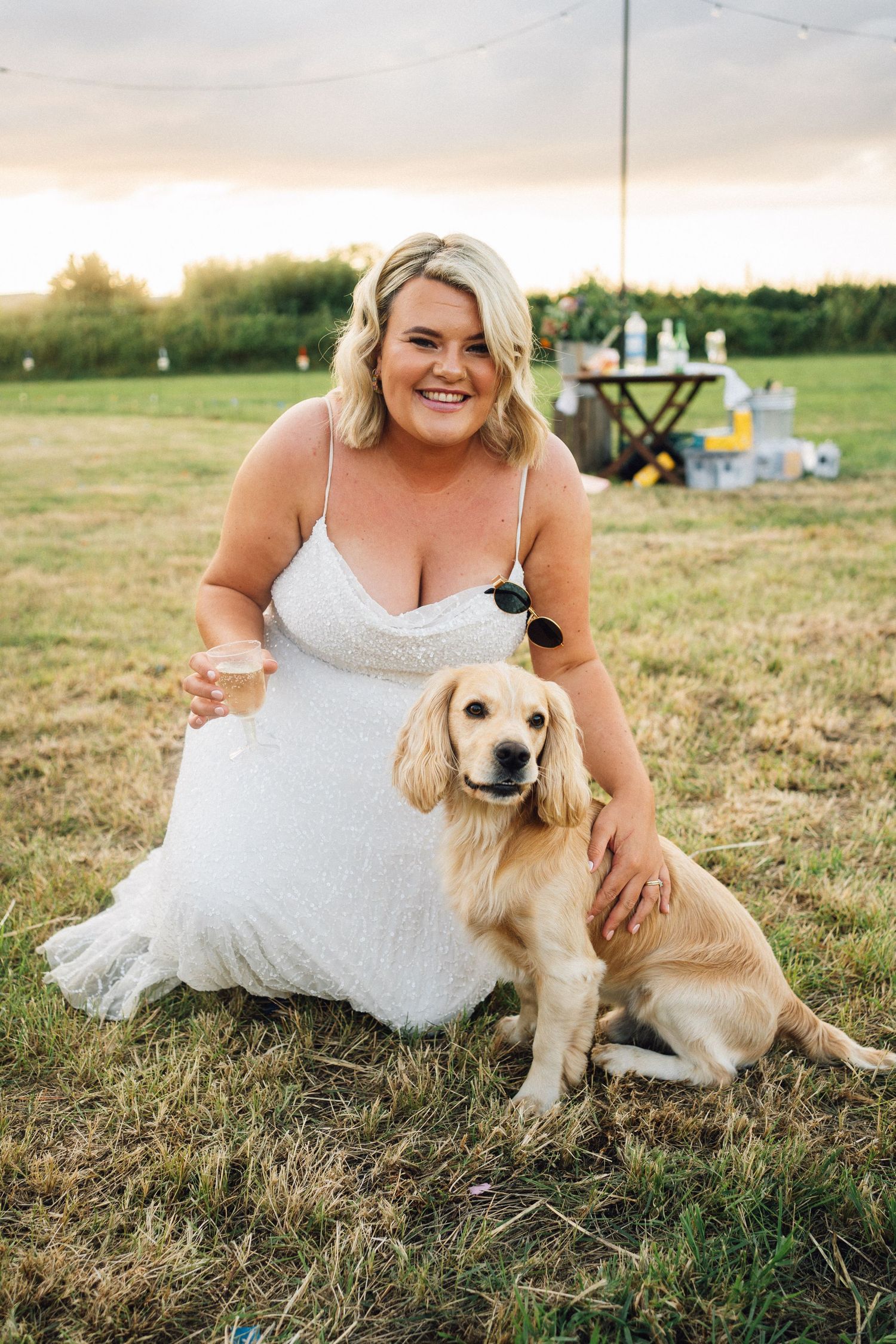 Golden retriever dog sits with person in white wedding dress on grassy field during sunset.
