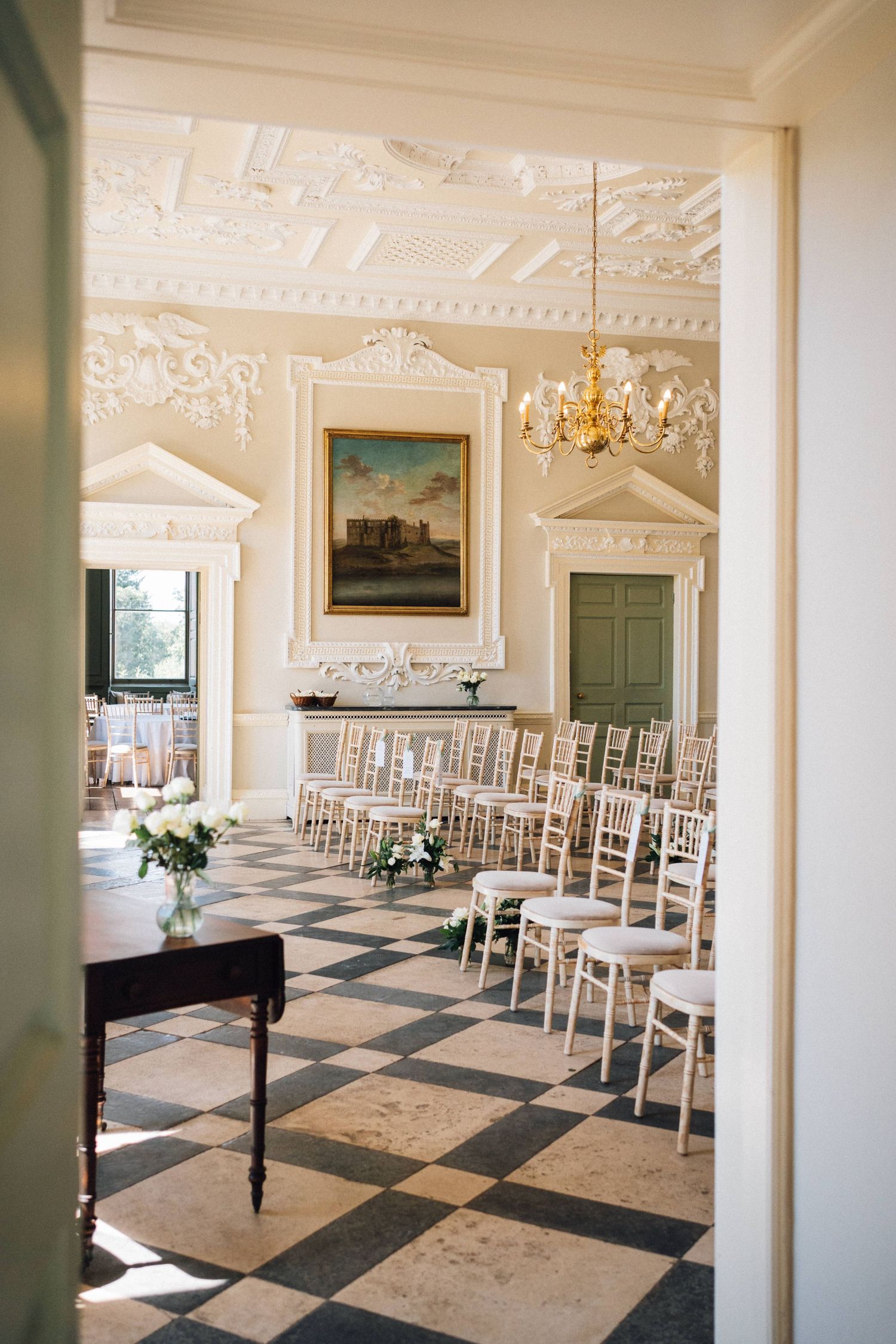 Elegant wedding ceremony venue with ornate white moldings, checkerboard floor and rows of white chairs.
