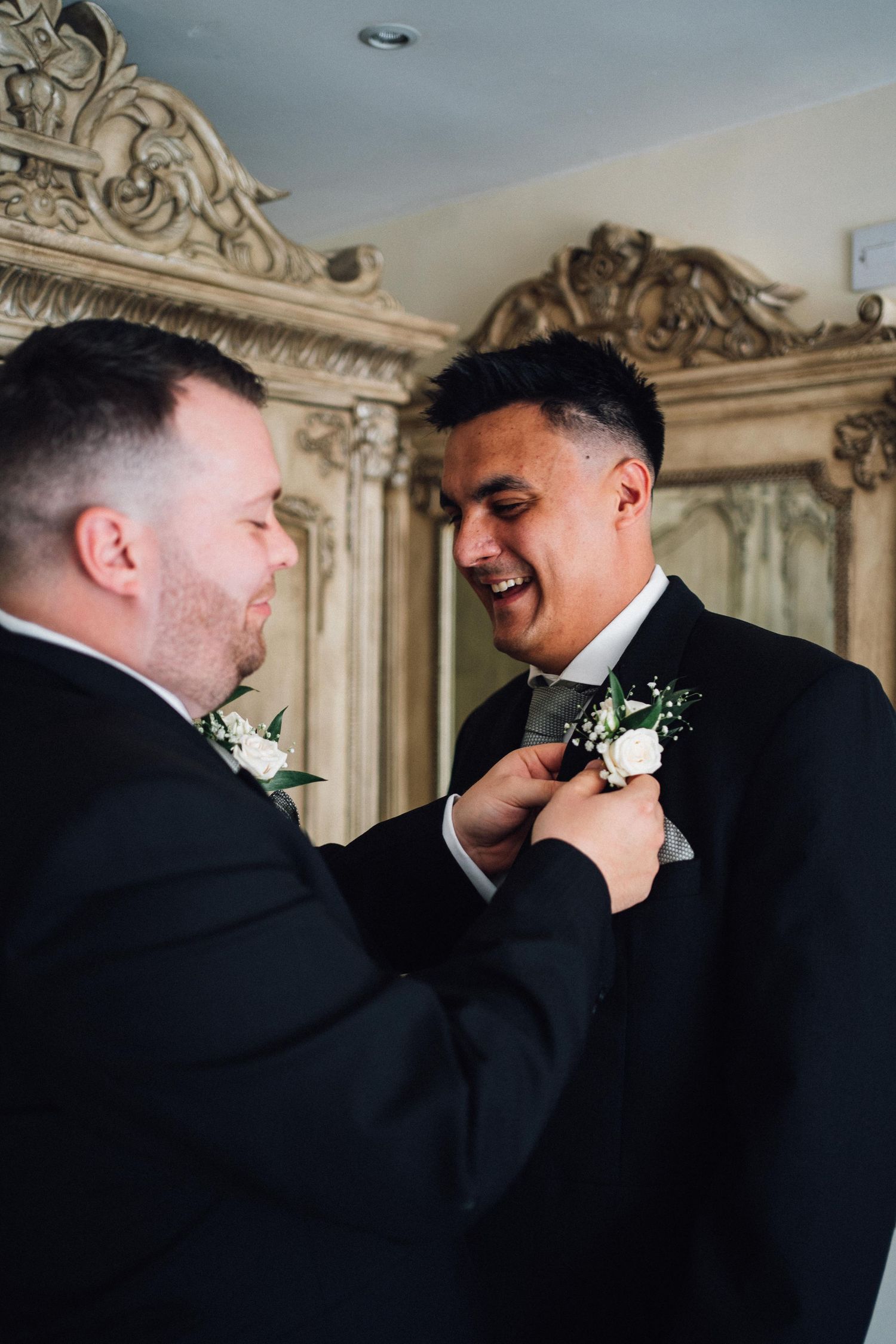 Two men in formal attire help each other with boutonnieres in an ornately decorated room.