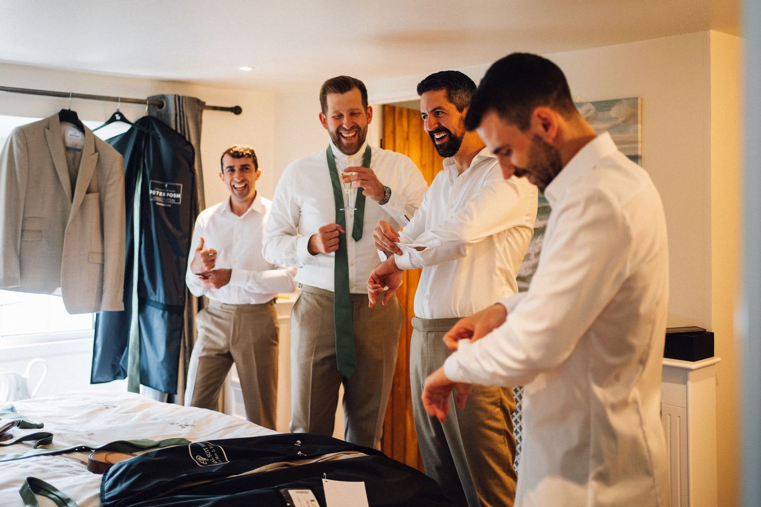 Groomsmen getting dressed and helping each other prepare in a bright hotel room before a wedding celebration.