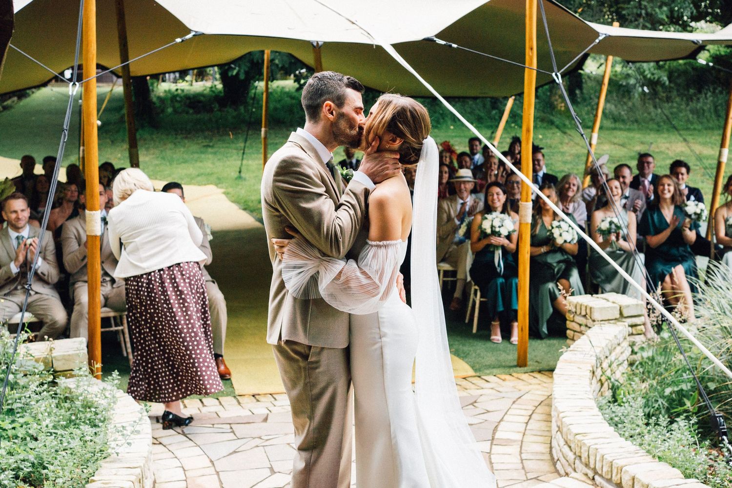 Wedding guests gather under a yellow canopy tent during an outdoor celebration in a garden setting.