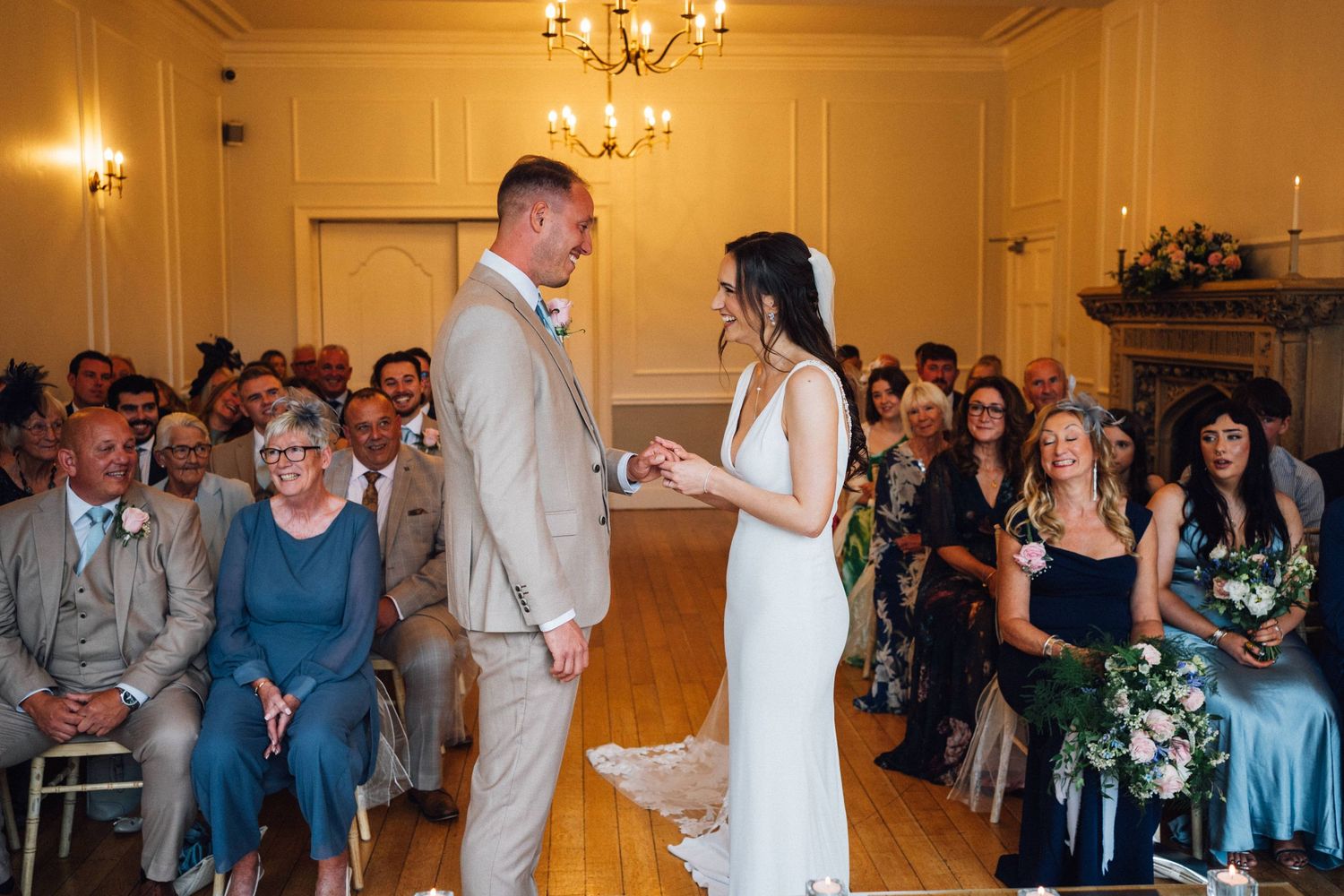 A couple exchanges vows during an indoor wedding ceremony with guests seated in rows and a chandelier overhead.