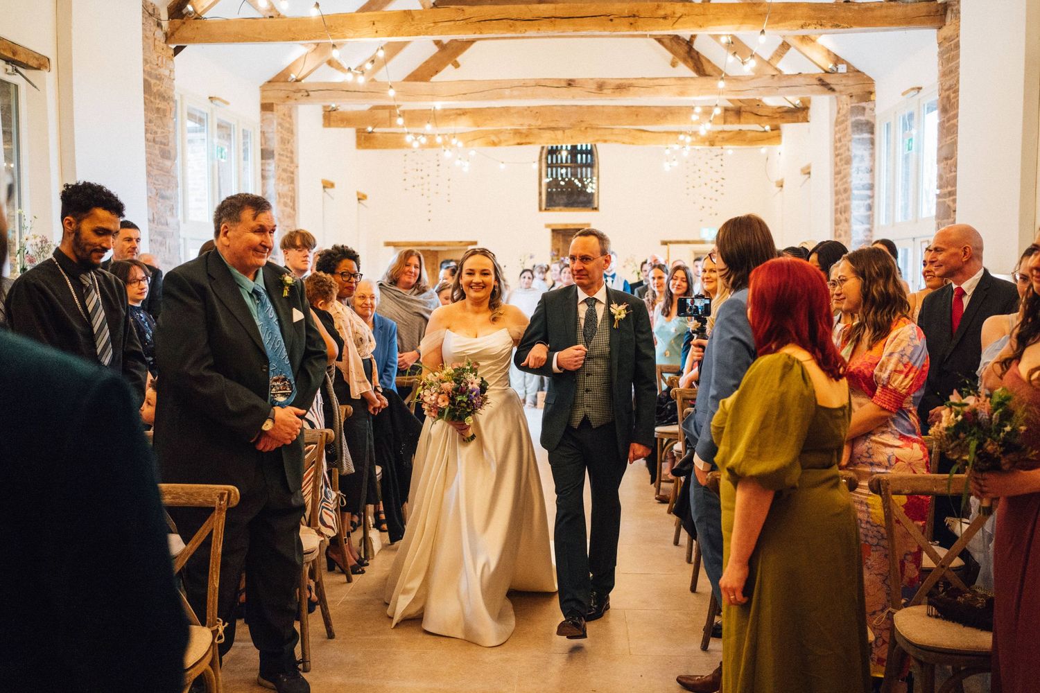 Wedding guests mingle and dance in a rustic barn venue with exposed wooden beams and warm lighting.