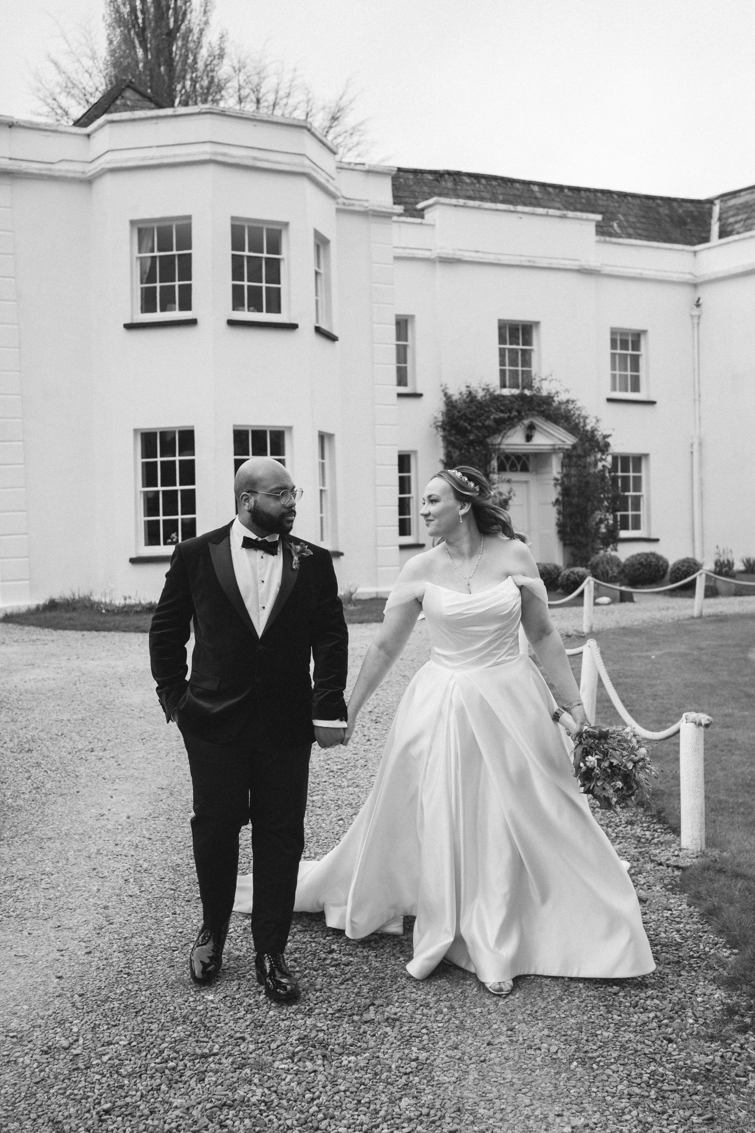 A black and white portrait of newlyweds walking together outside an elegant white country house.