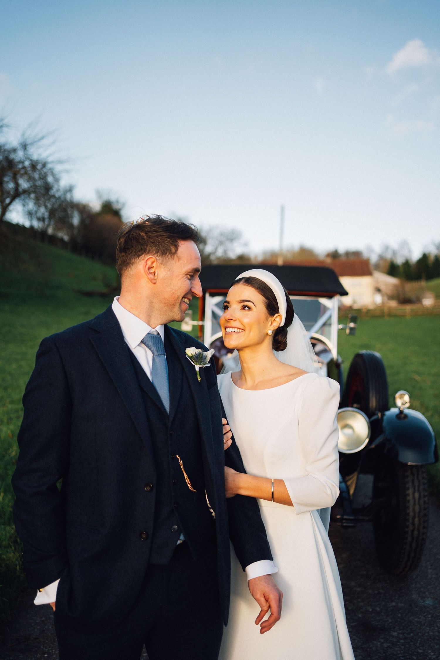 A romantic wedding photo of a couple sharing a joyful moment together next to a vintage car in a countryside setting.