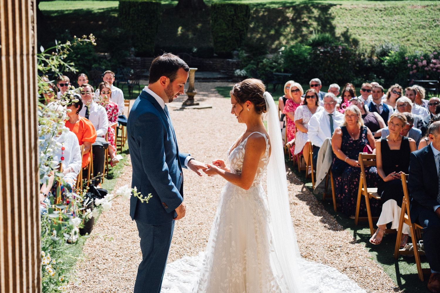 Couple exchanging vows at an outdoor wedding ceremony with guests seated along a gravel aisle.