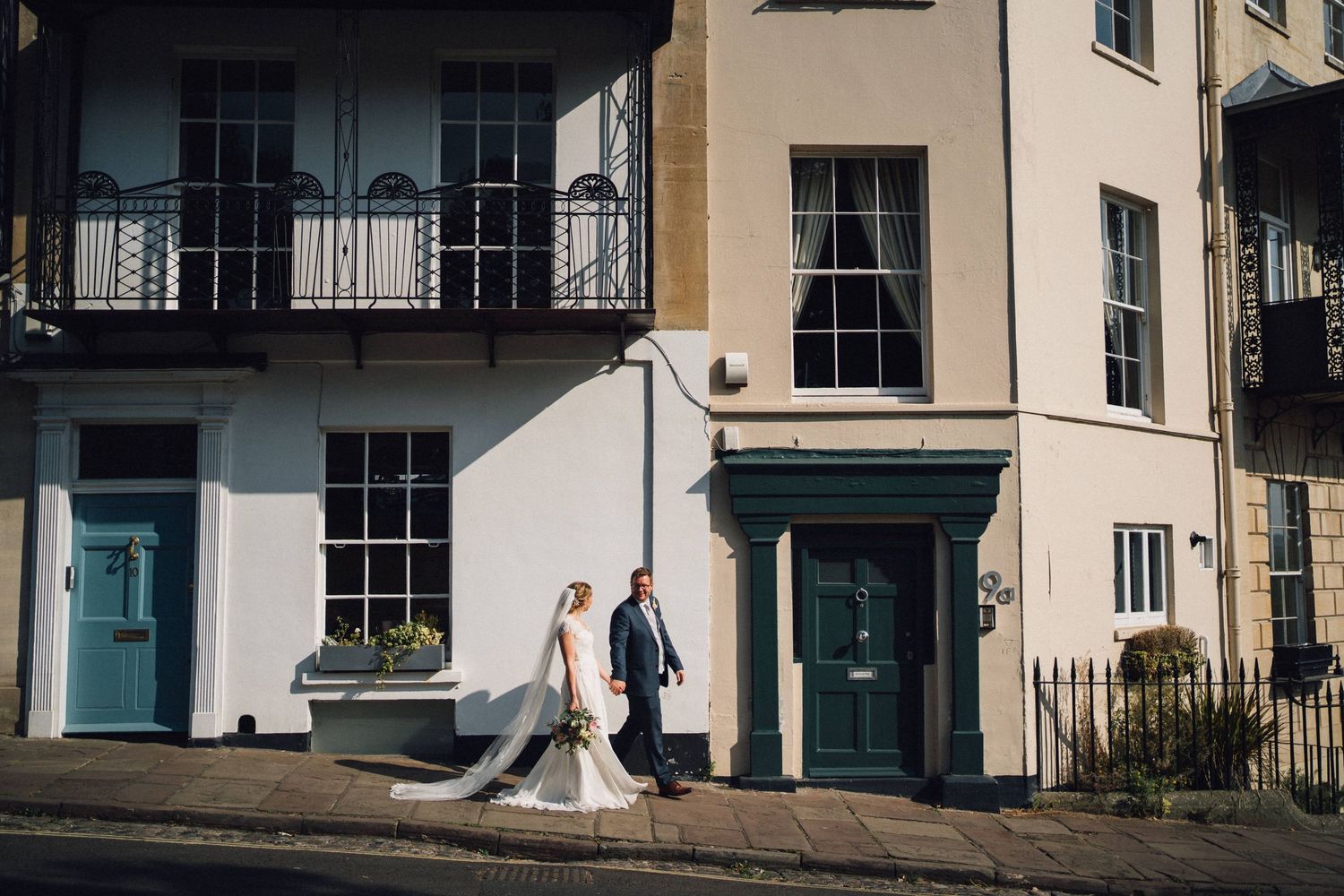 Bride and groom walking outside elegant cream Georgian building with teal doors on sunny wedding day.