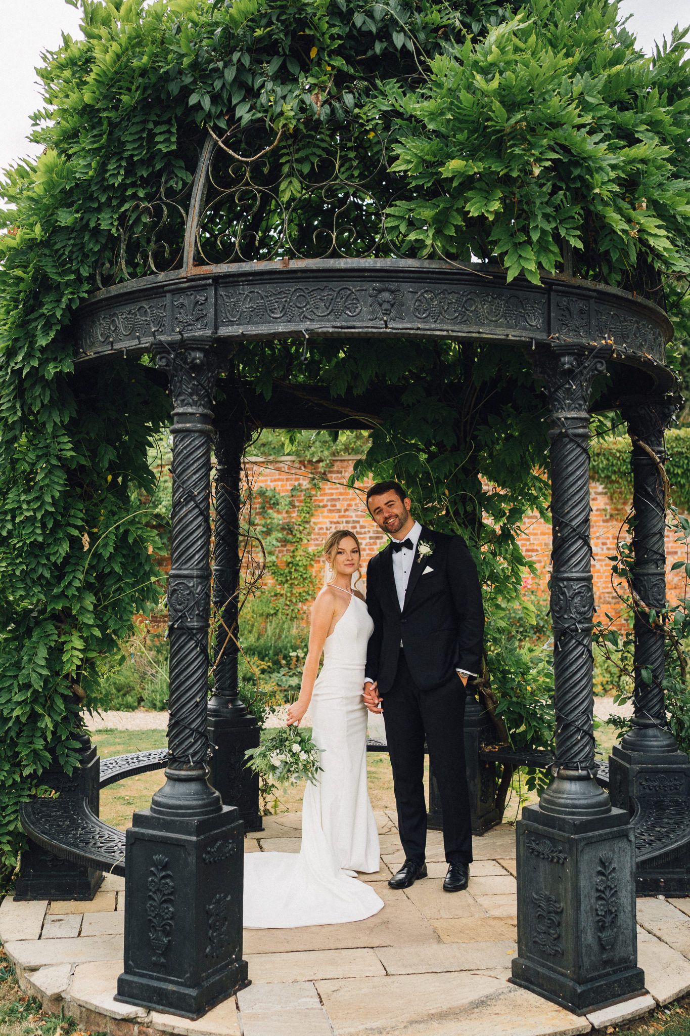 Newlyweds share a moment under a decorative black iron gazebo in a garden setting.